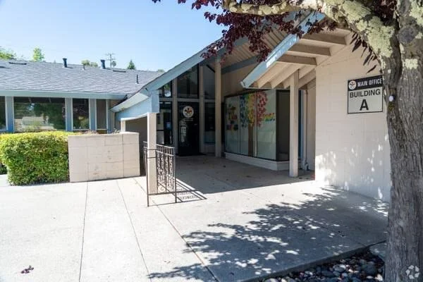 Entrance to a school building with a sign reading 'Main Office, Building A', large glass windows, and a tree casting shade on the sidewalk.