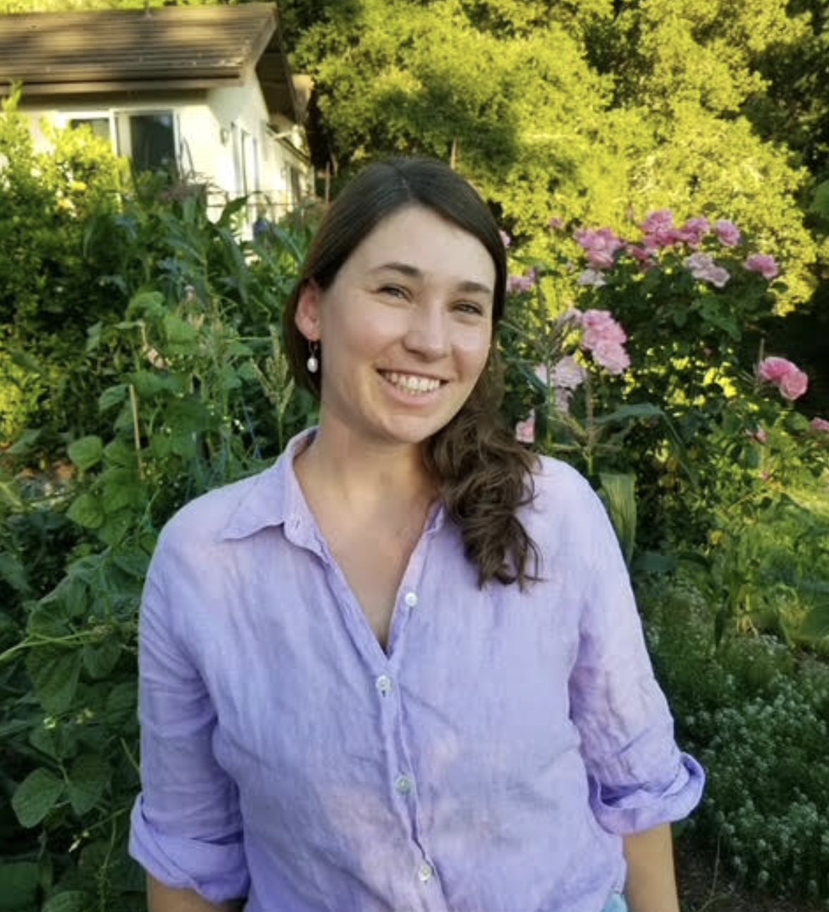 A woman smiling outdoors surrounded by green foliage and pink flowers, wearing a light purple shirt and pearl earrings.