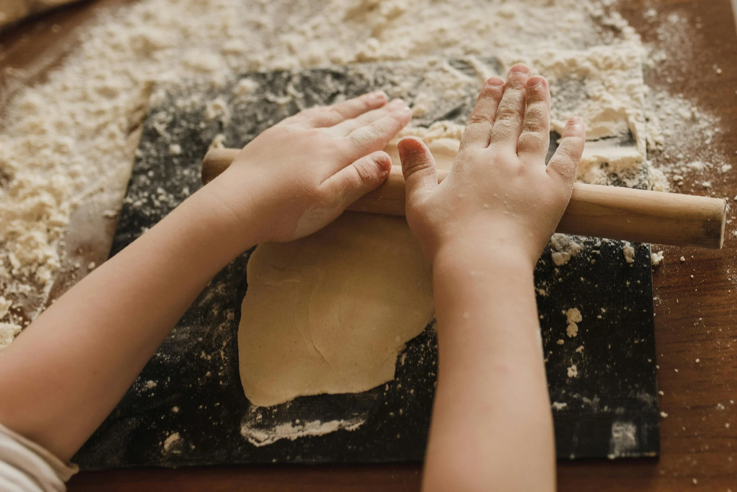 Two young children's hands rolling out dough with a rolling pin on a floured surface.