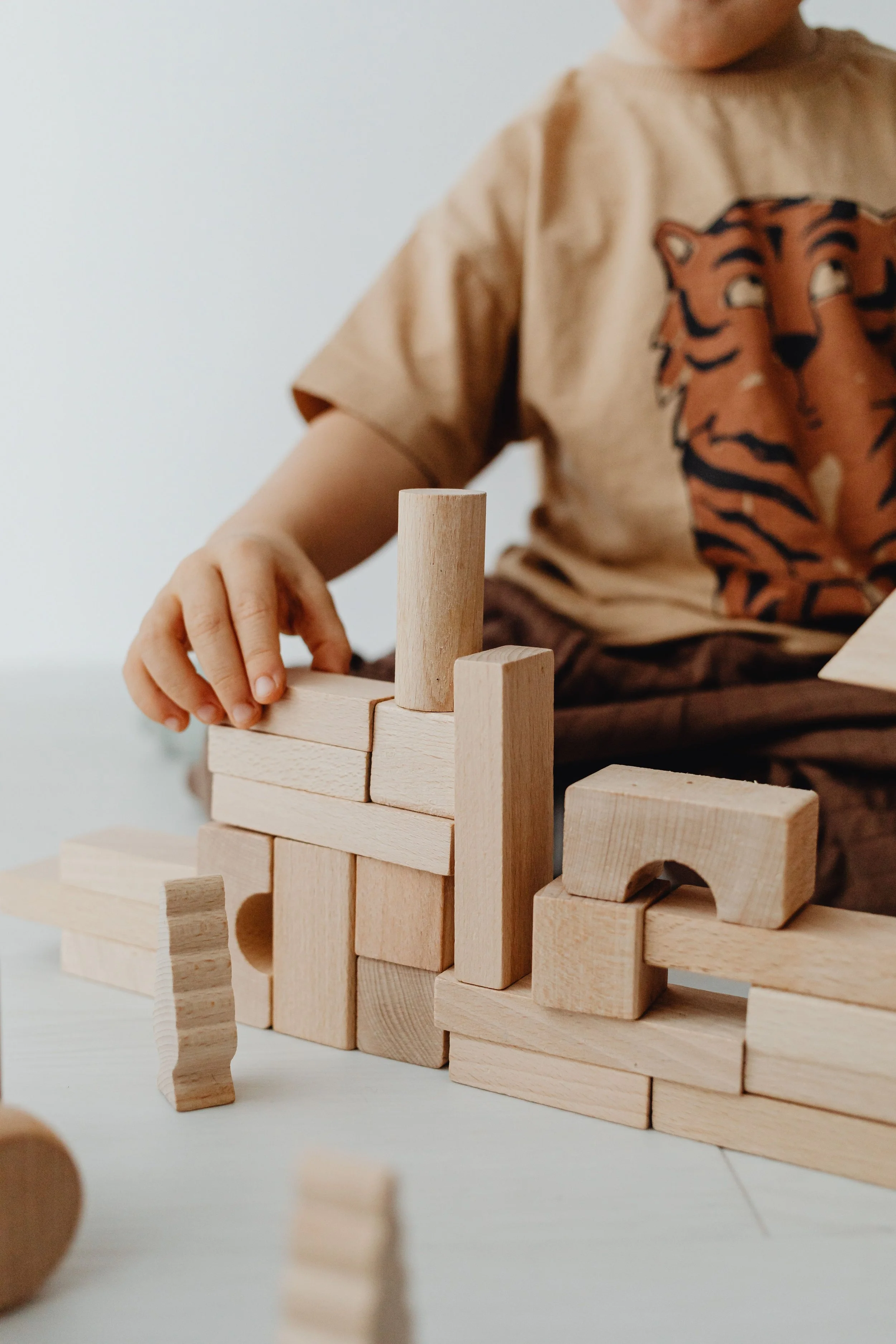 A child wearing a beige T-shirt with a tiger face print is playing with wooden blocks, stacking and arranging them on a white surface.