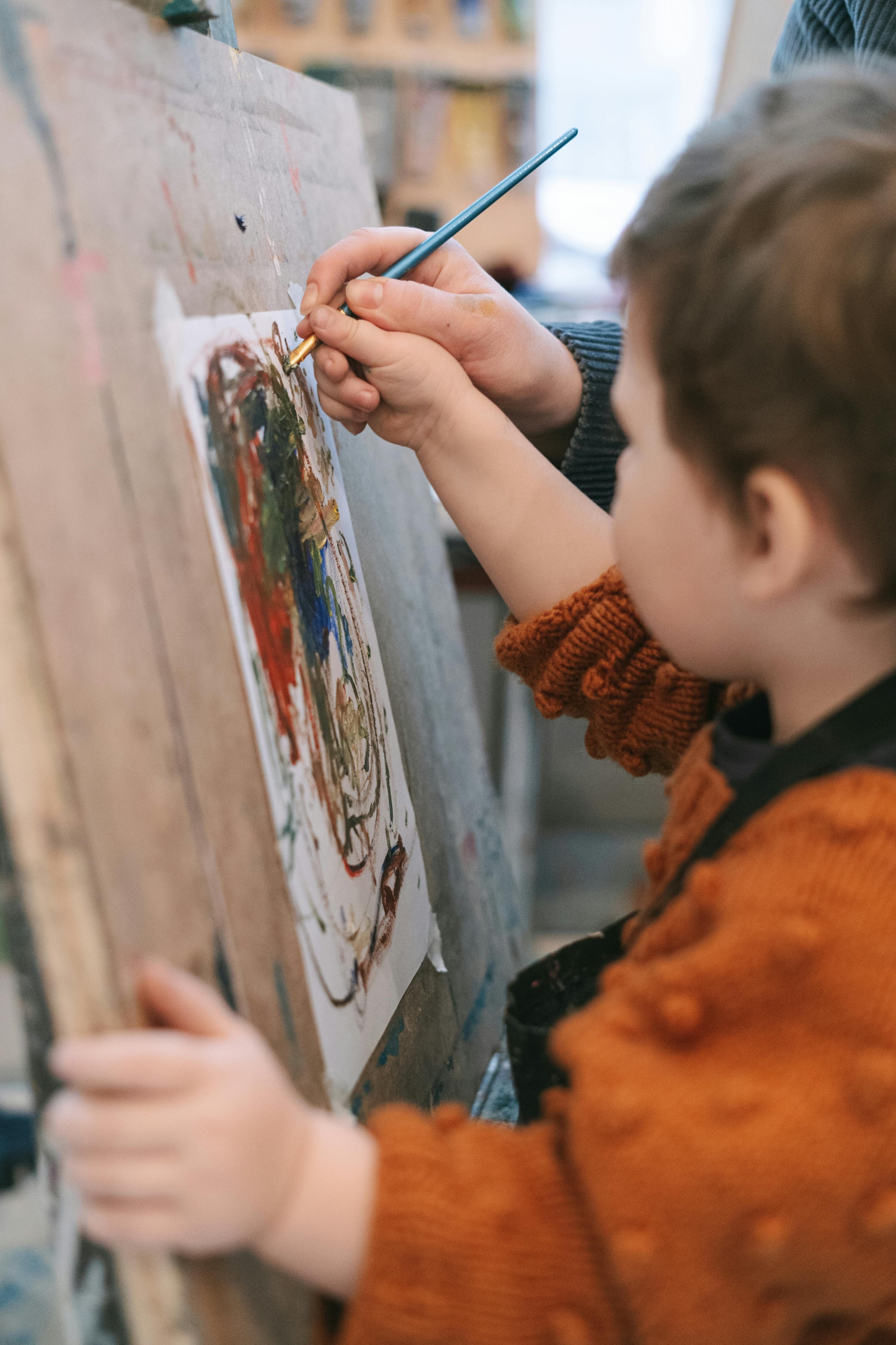 A child with brown hair and an orange sweater painting on a canvas with an adult helper using a thin brush.