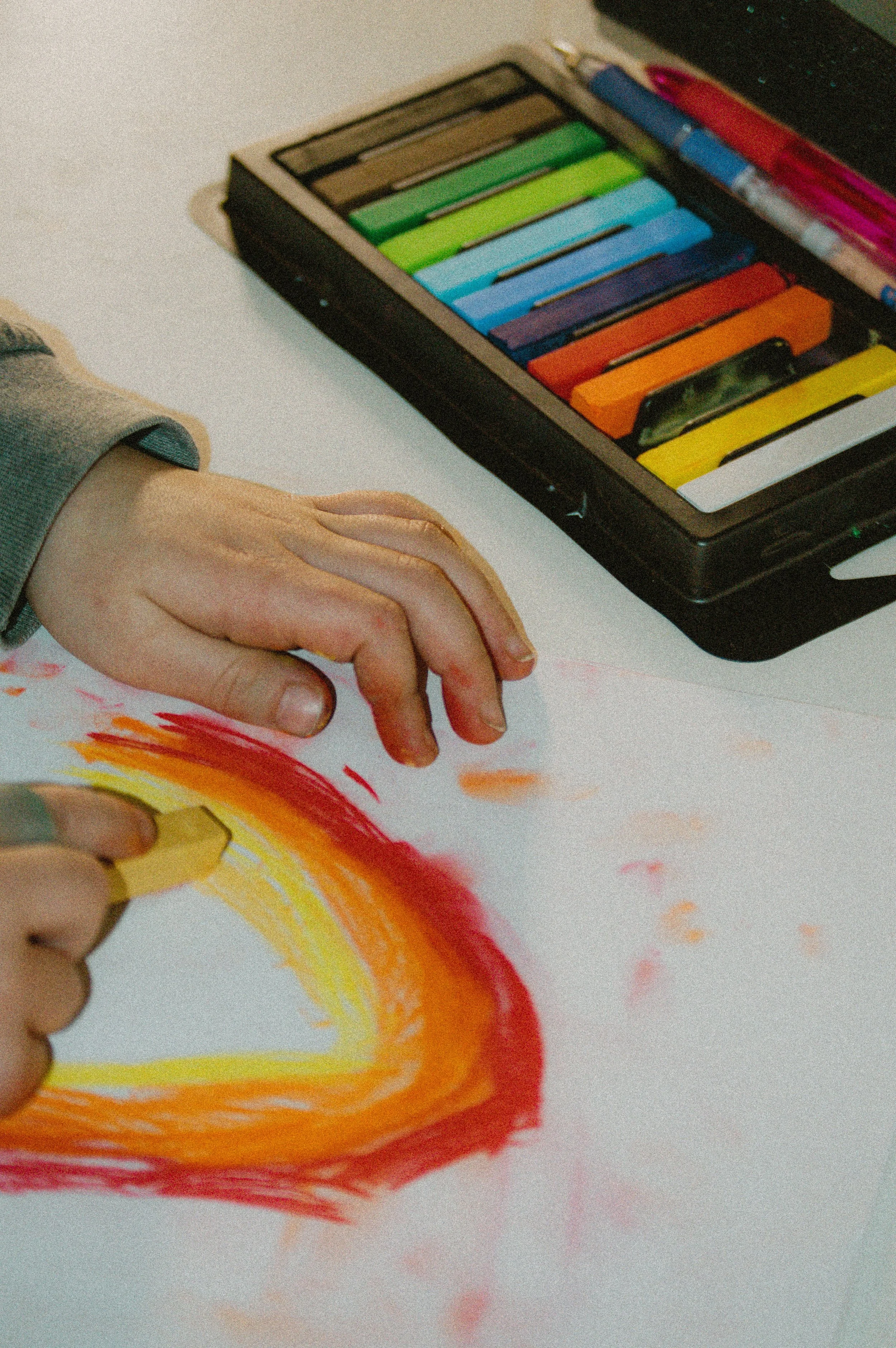 Child's hand drawing a rainbow with colorful markers on paper, with a case of various colored markers beside.