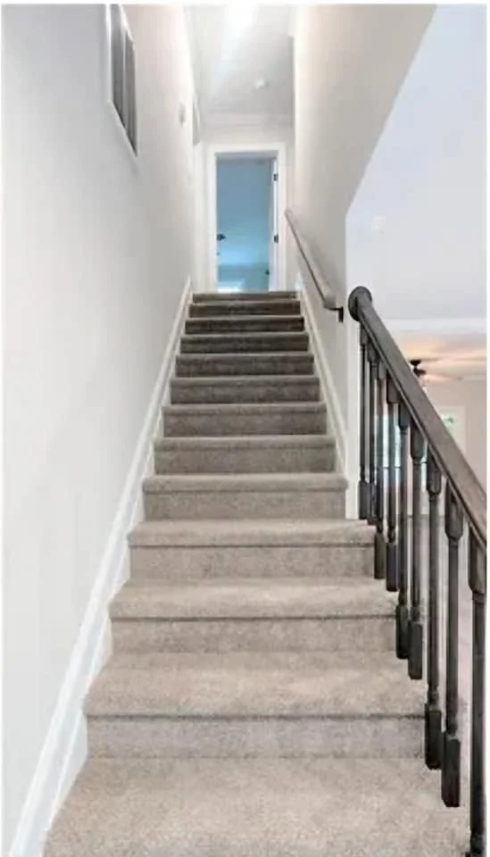 Interior view of a staircase with carpeted steps, beige walls, and a wooden and metal handrail on the right side, leading up to a door at the top.