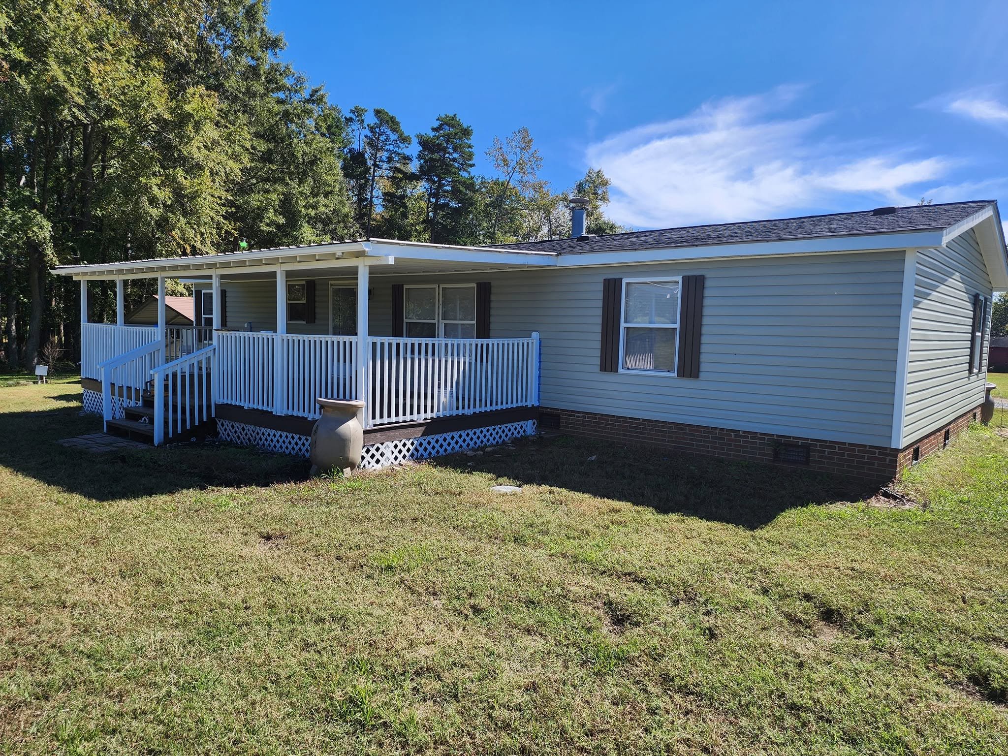 The image shows a house with a light gray exterior, white trim, and dark brown shutters. The house has a small front porch with white railings and steps. It is surrounded by a green lawn with some small shrubs, and there are tall trees in the backgro