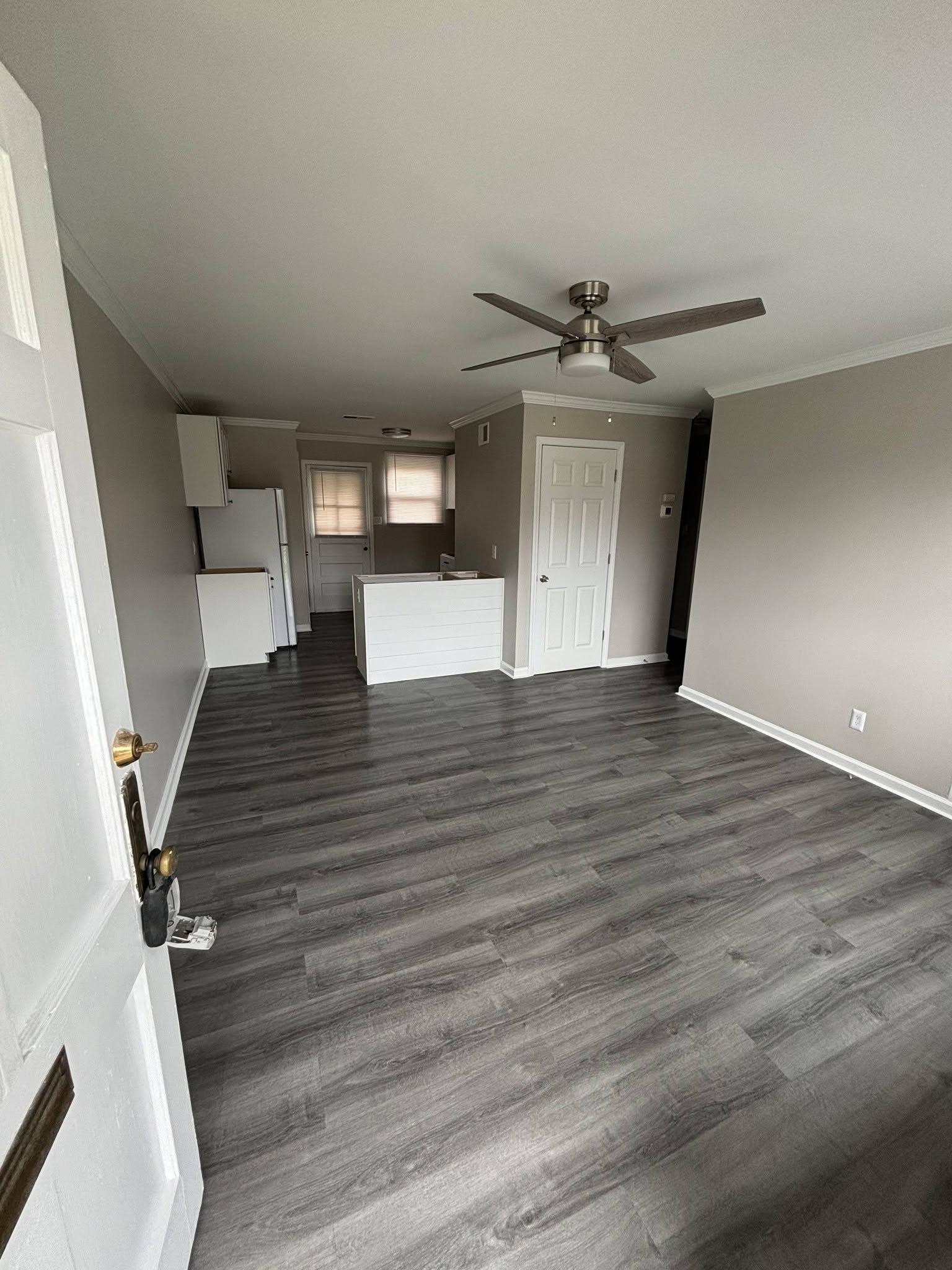 Empty living room and kitchen with wood-style flooring, ceiling fan, white cabinetry, gray walls, and a door with windows.