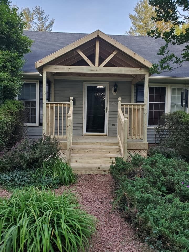 House entrance with a small wooden porch, newly built steps, and a gabled roof under construction. Surrounded by greenery and bushes.