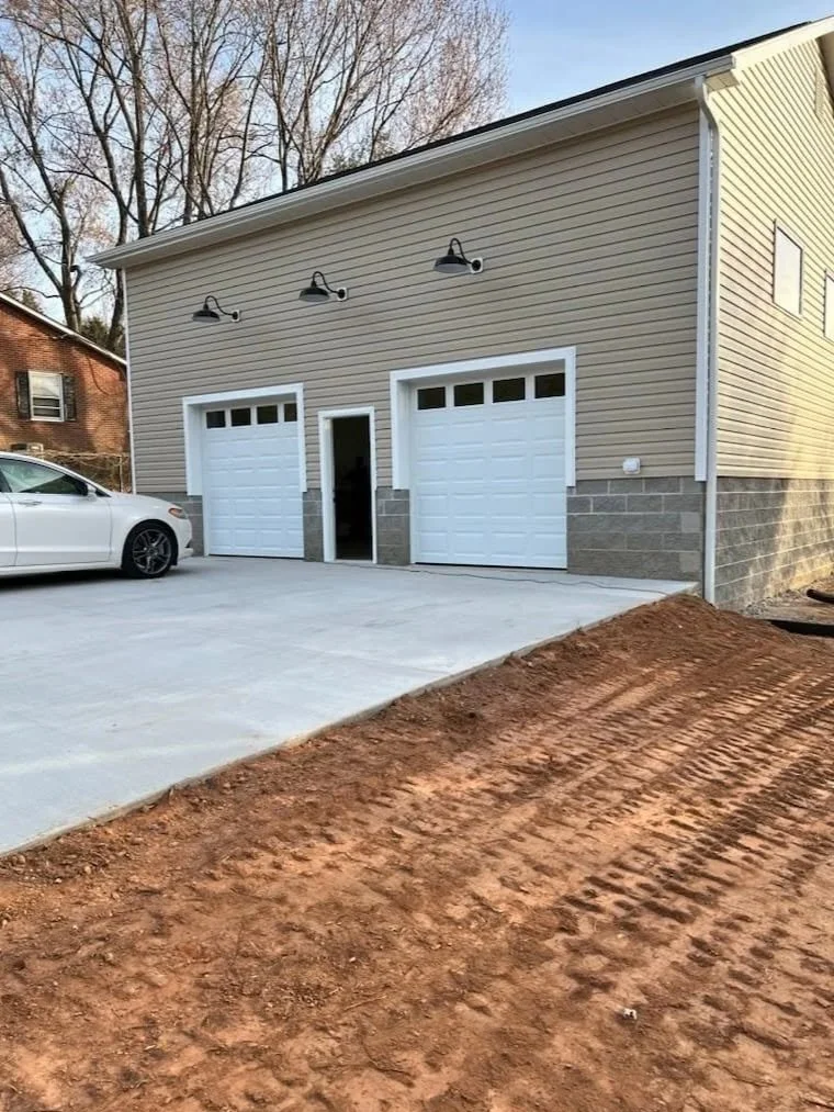 any property renovations Newly paved concrete driveway leading to a garage with two doors and a small door in the middle, attached to a beige building with three outdoor lights, a white car parked on the driveway, and brown dirt in the foreground.