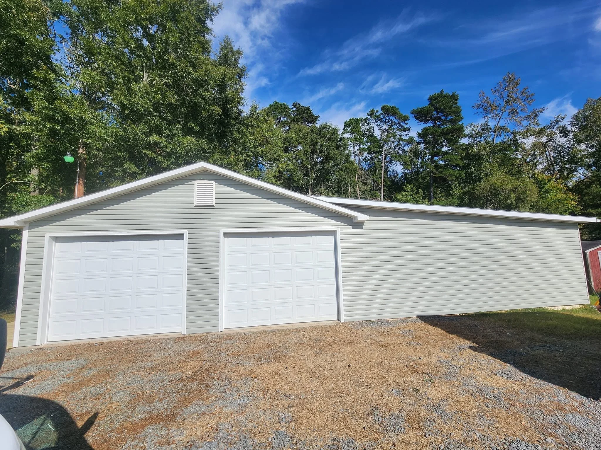 any property renovations A light gray garage with two closed doors and a gabled roof, situated in front of a wooded area with tall trees, under a blue sky with scattered clouds.
