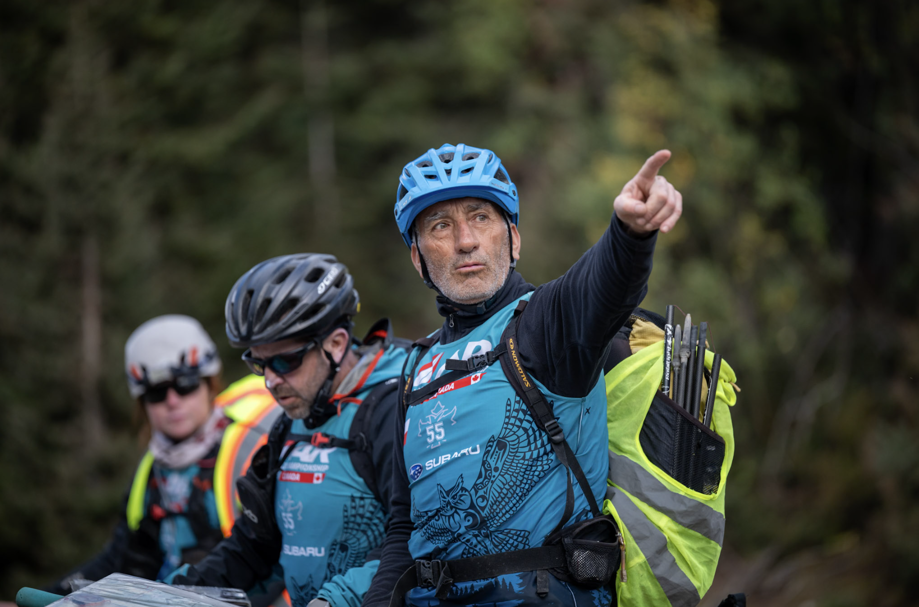 A group of Adventure Racers in helmets and outdoor gear, with one man pointing forward, in a forested area.