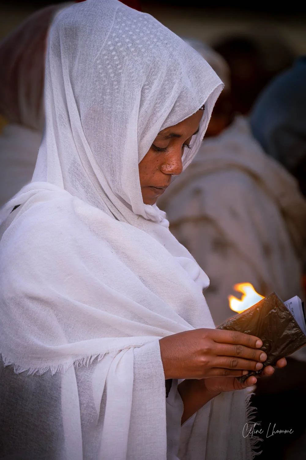 A woman dressed in white robes and a headscarf holding a small object with a flame, possibly during a religious or spiritual ceremony.