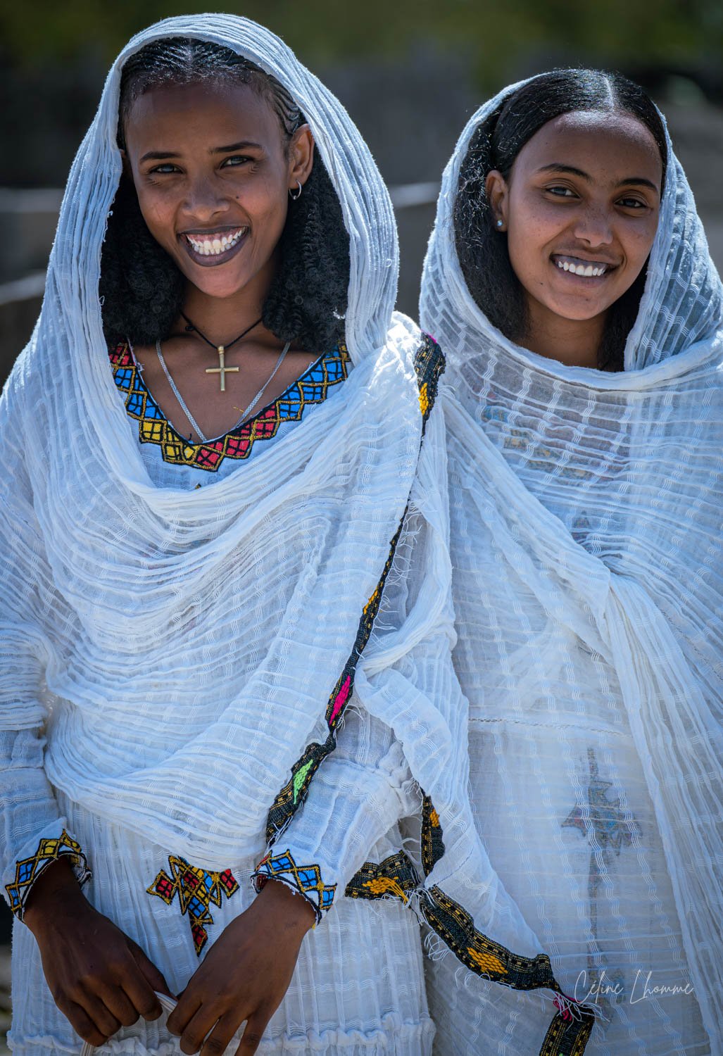 Two women wearing traditional white attire with colorful embroidery, smiling outdoors.
