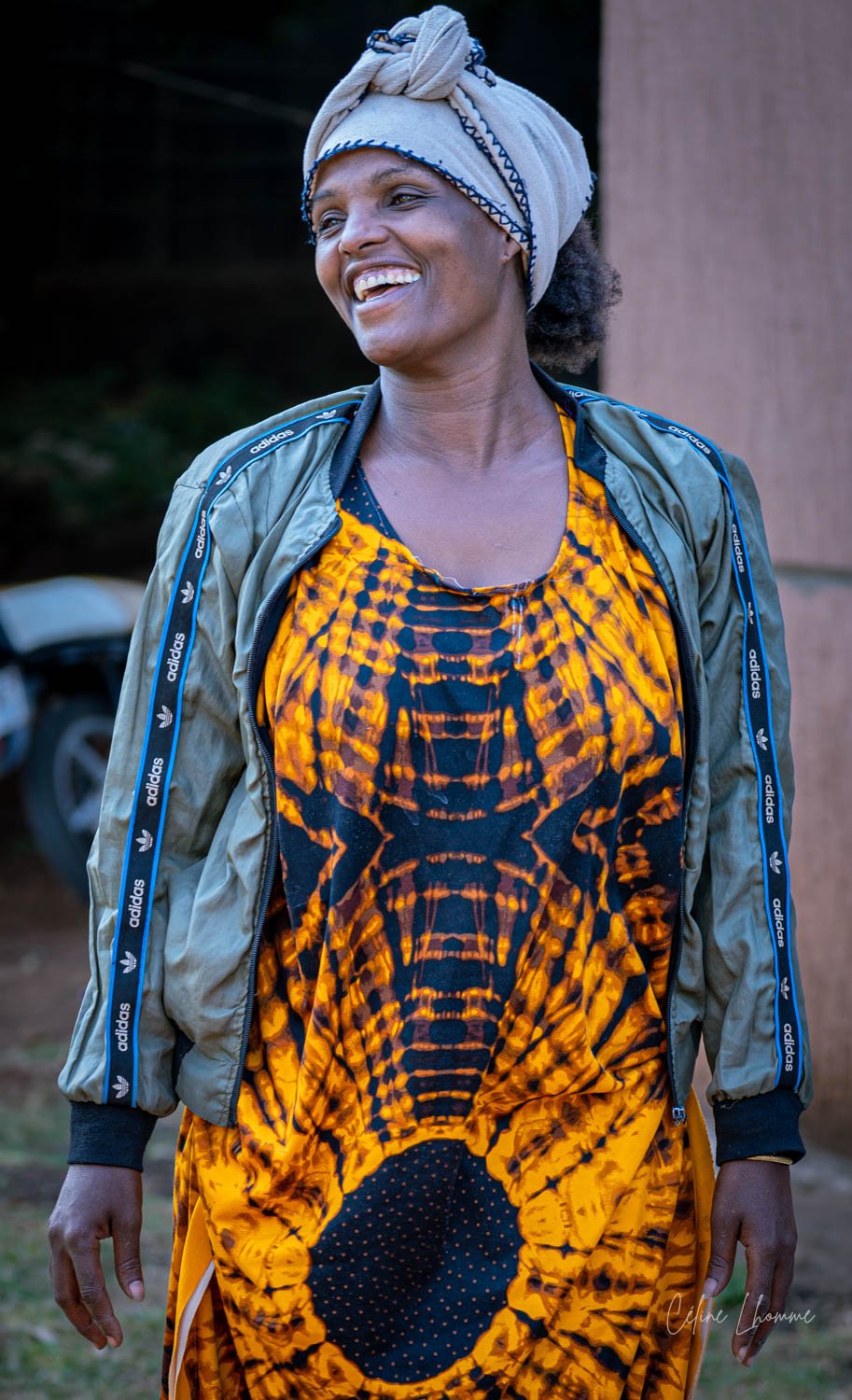 Smiling woman wearing a beige headwrap, a green Adidas jacket with blue stripes, and a yellow and black patterned dress, outdoors.