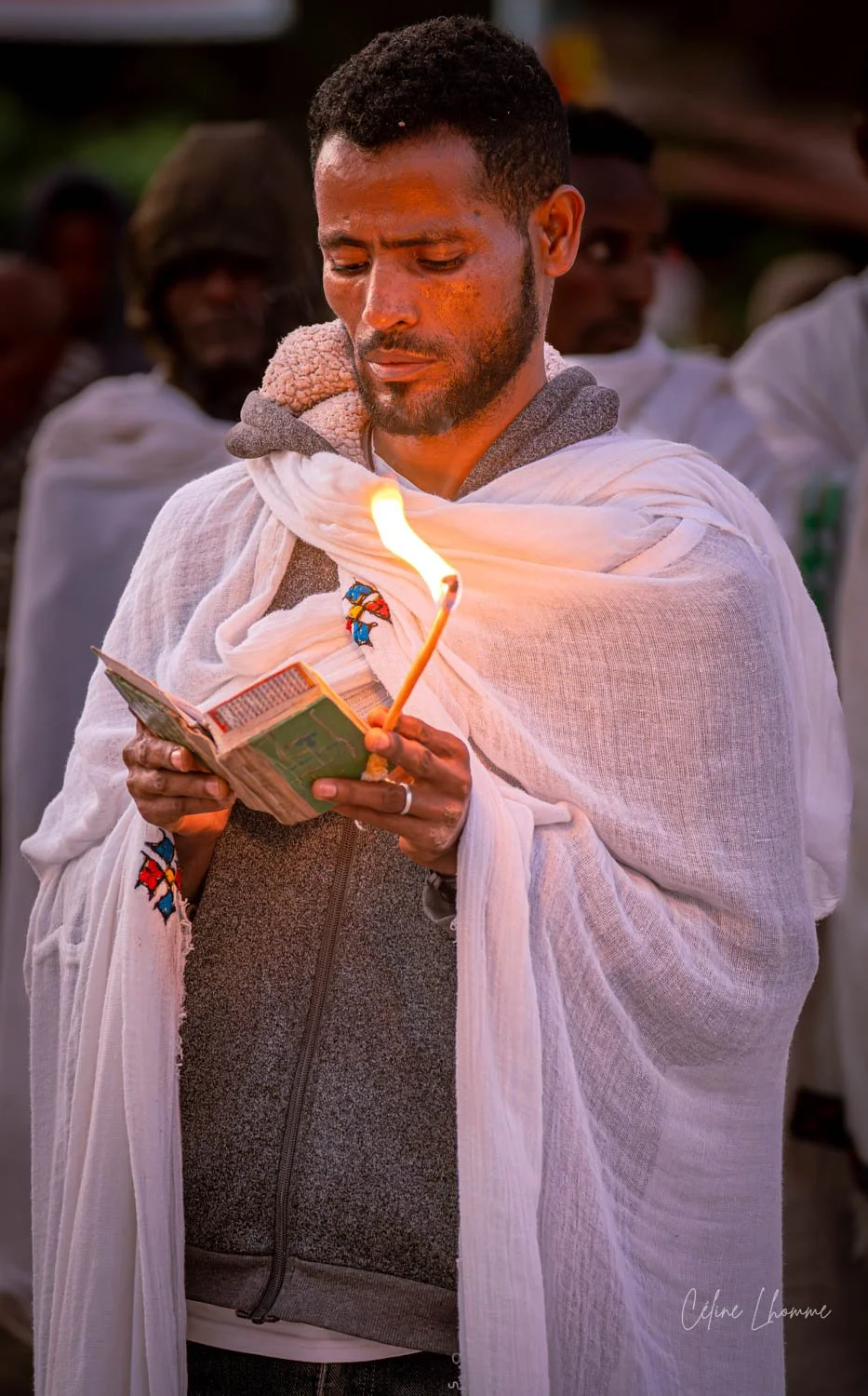 A man holding a lit candle, reading a prayer book, wrapped in a white shawl, during a religious ceremony or prayer gathering.