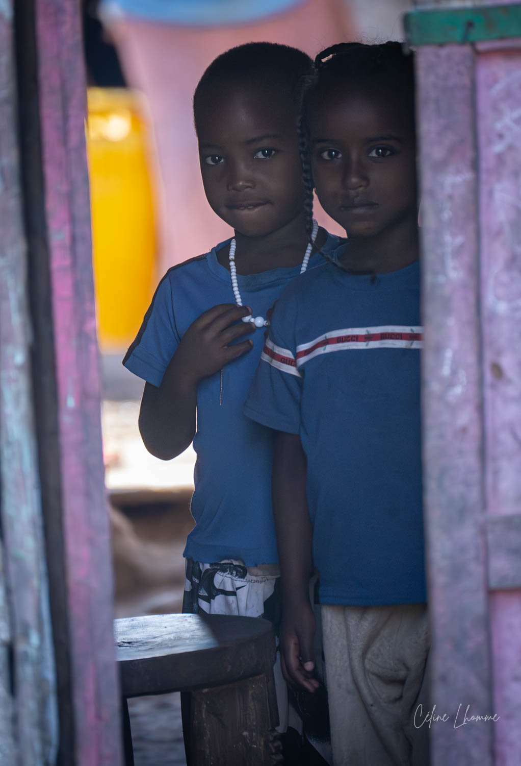 Two young girls with dark skin and braided hair peeking through a doorway, wearing blue shirts, with a pink wall and yellow light in the background.