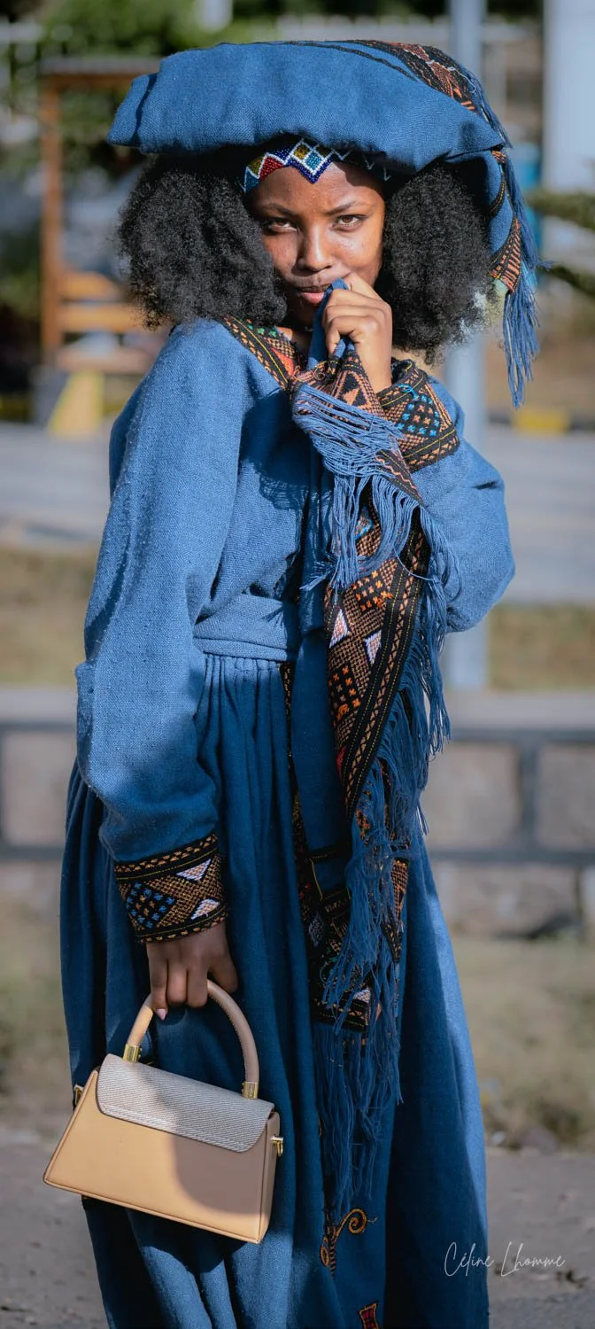 Woman outdoors in a blue traditional or cultural outfit, wearing matching headgear, holding a beige handbag, with curly hair and a thoughtful expression.