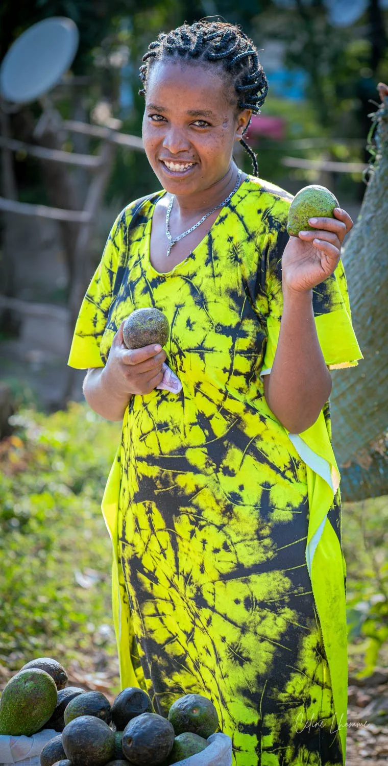 Smiling woman in yellow and black patterned dress holding green avocados outdoors with a basket of more avocados in front of her.