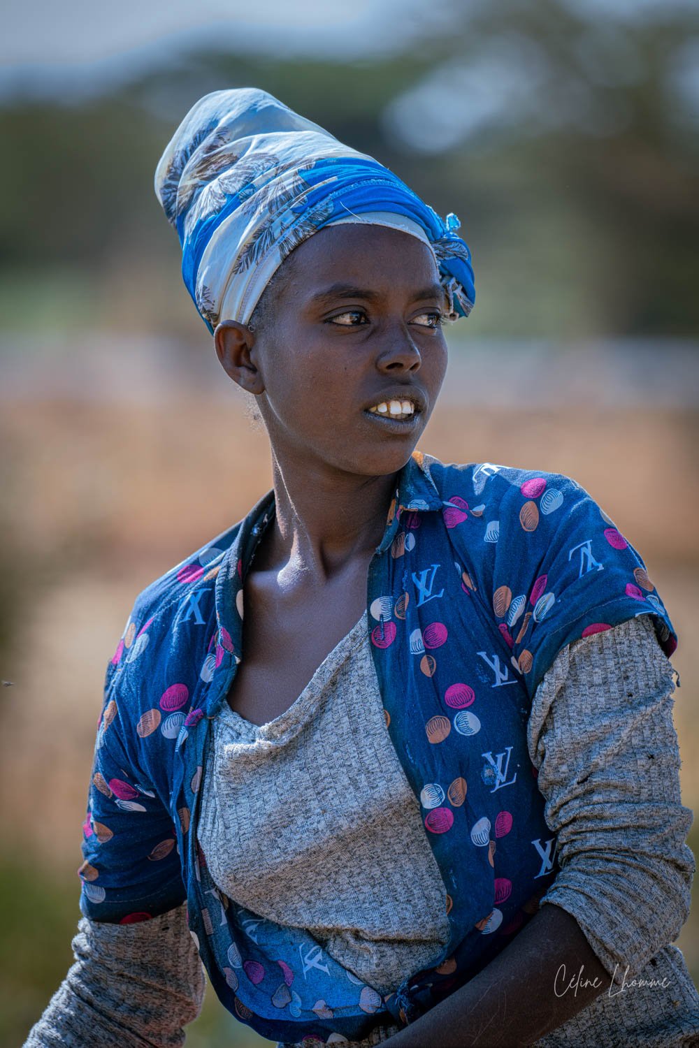 A young woman with dark skin is wearing a blue patterned shirt and a headscarf with shades of blue and white, outdoors with a blurred background.
