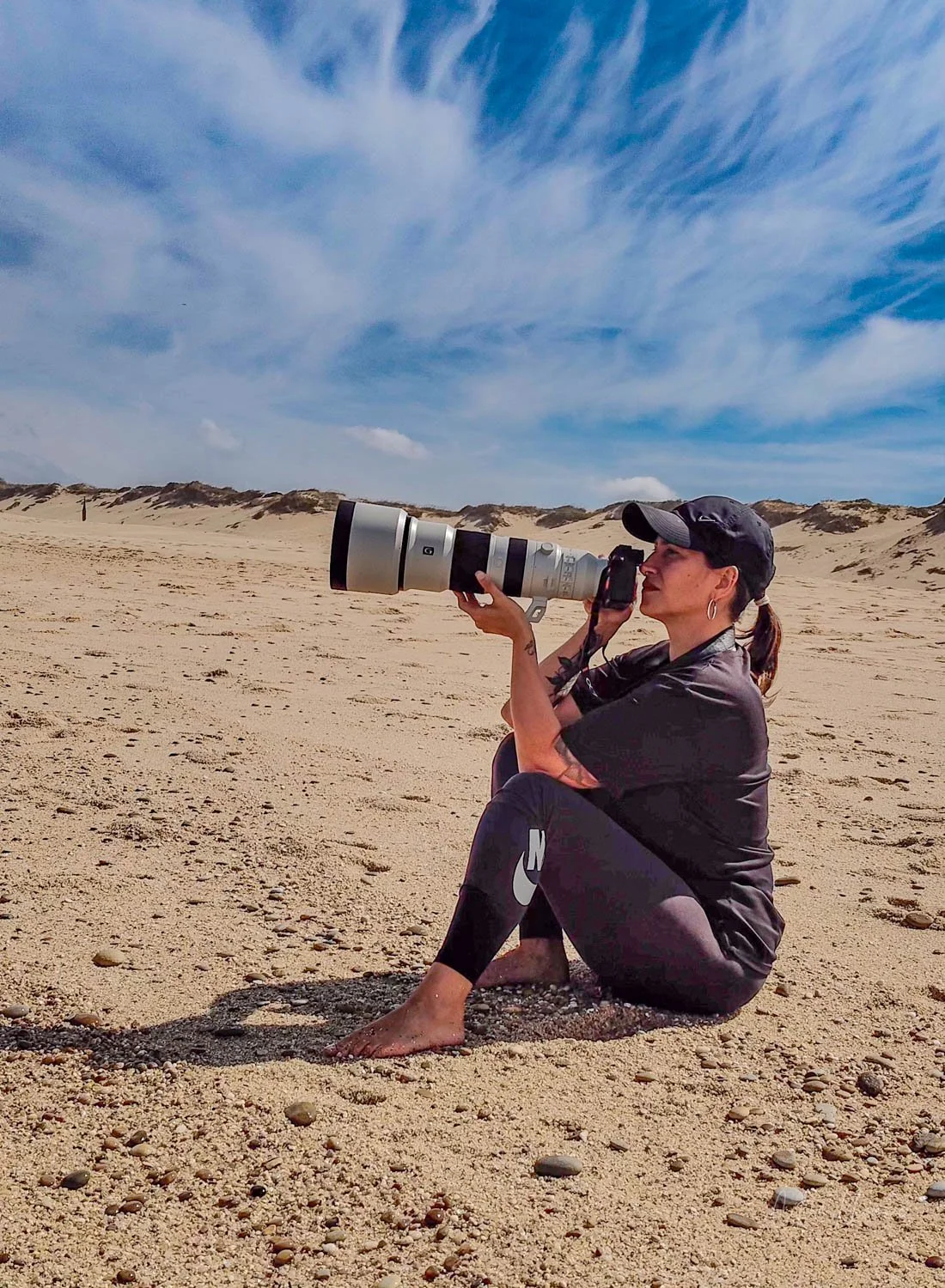 A woman sitting on the sandy beach, holding a large camera with a telephoto lens, wearing a black cap, earrings, and black athletic clothing, with beach dunes and a cloudy sky in the background.