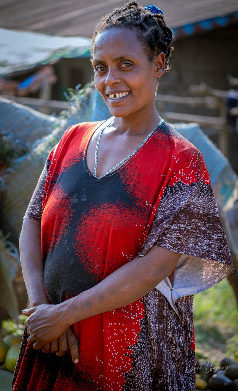 A smiling woman with dark skin and short, curly hair, wearing a colorful red, black, and brown patterned dress, stands outdoors with blurred structures and greenery in the background.