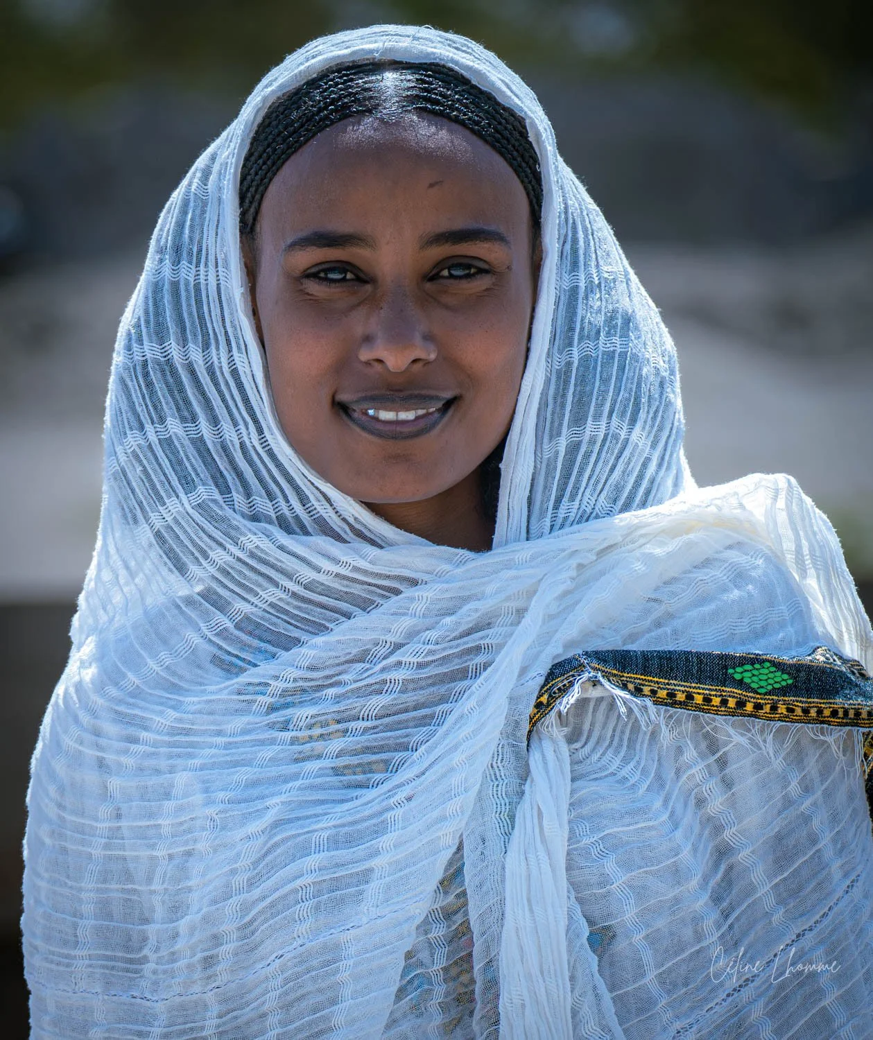 A woman with dark skin and braided hair, wearing a white patterned shawl, smiling outdoors.