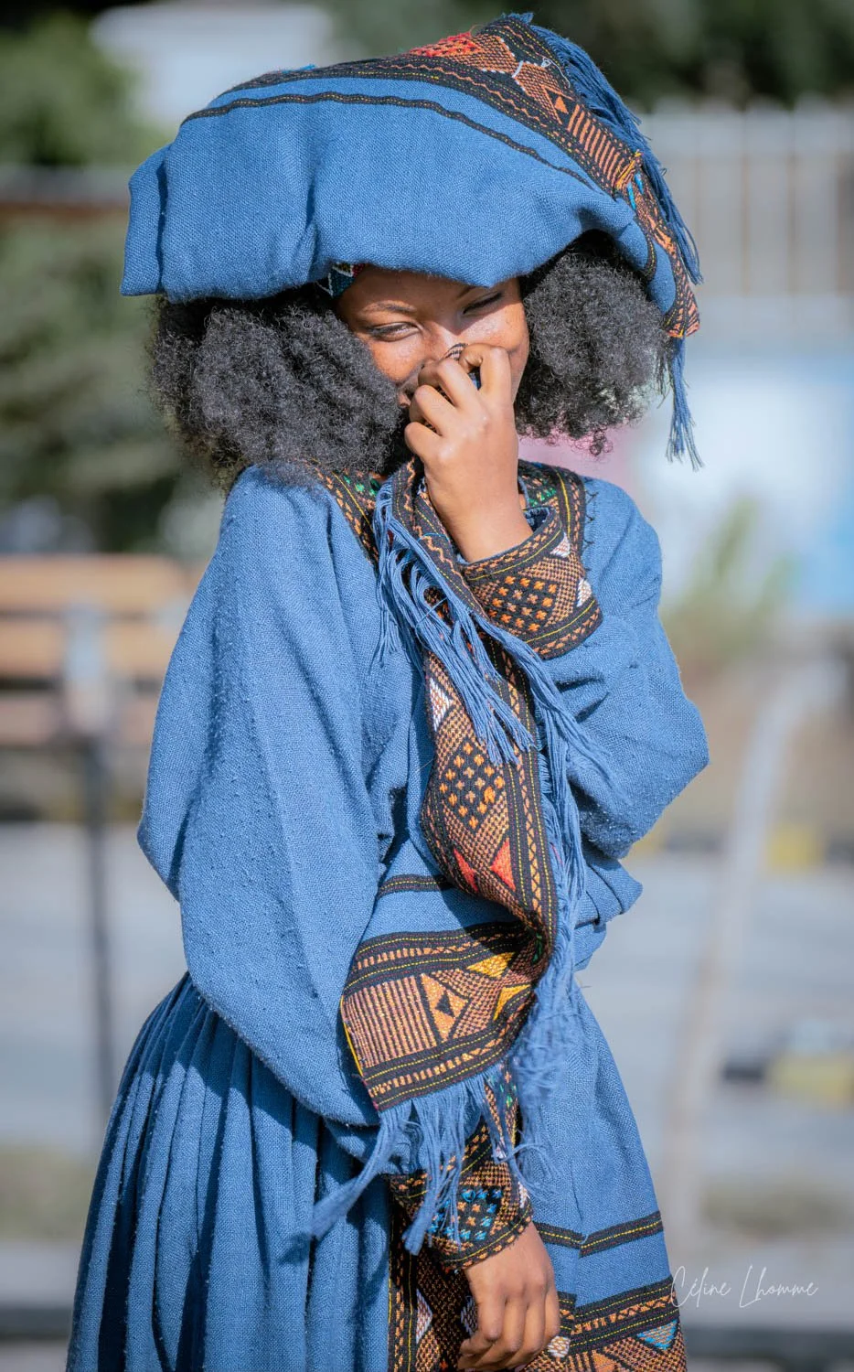 A woman wearing traditional African clothing and a large blue headscarf, smiling and covering her mouth with her hand.