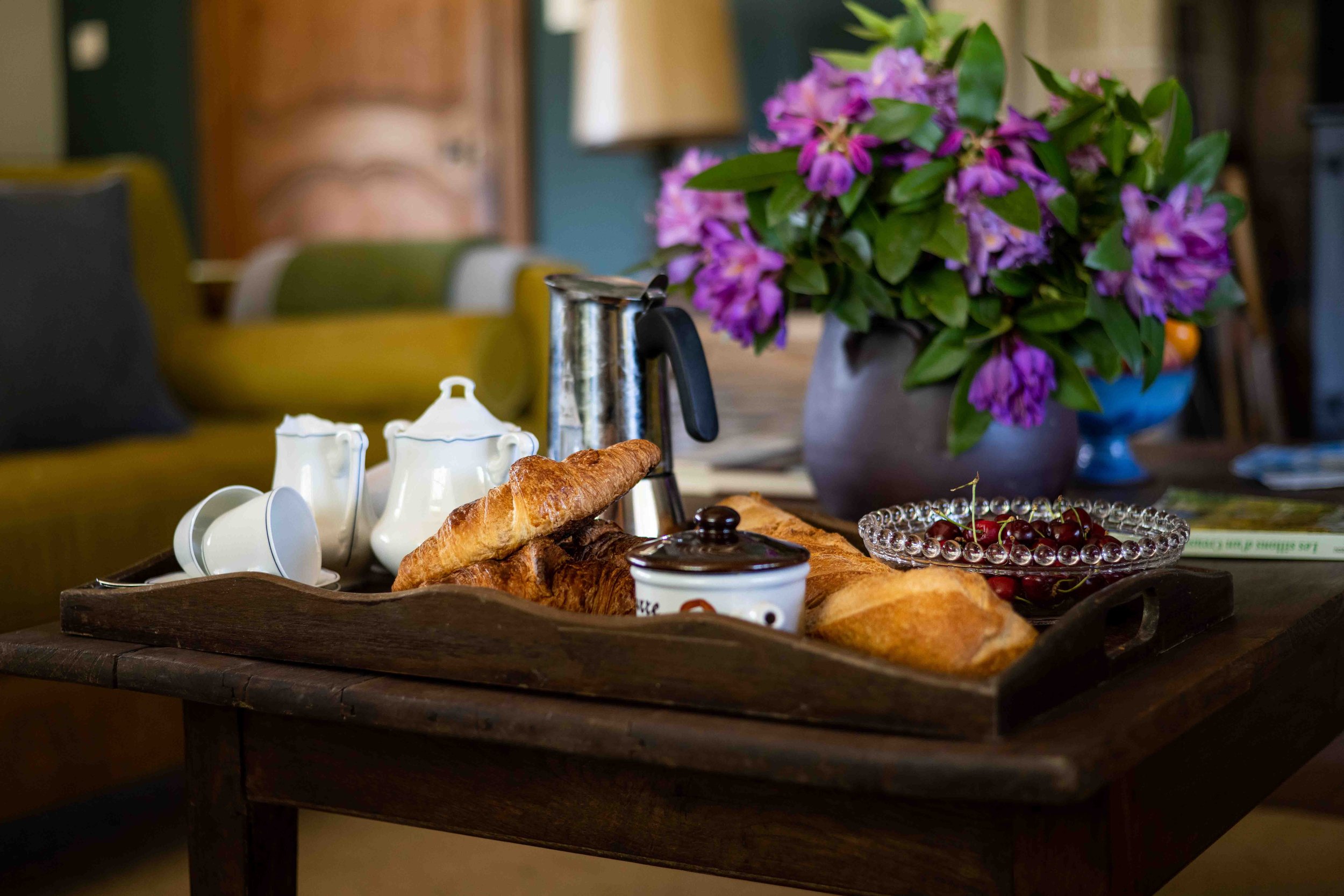 A wooden table set with breakfast items including croissants, a bowl of cherries, a teapot, cups, and a small jar, with a large purple floral arrangement in a gray vase in the background.