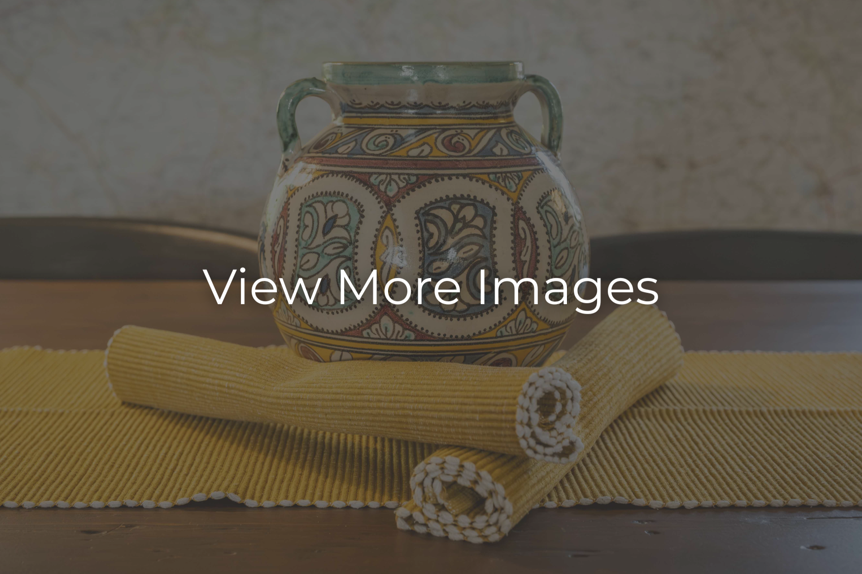 Decorative ceramic vase with intricate patterns on a yellow placemat, accompanied by a rolled yellow cloth with white accents, on a wooden surface.