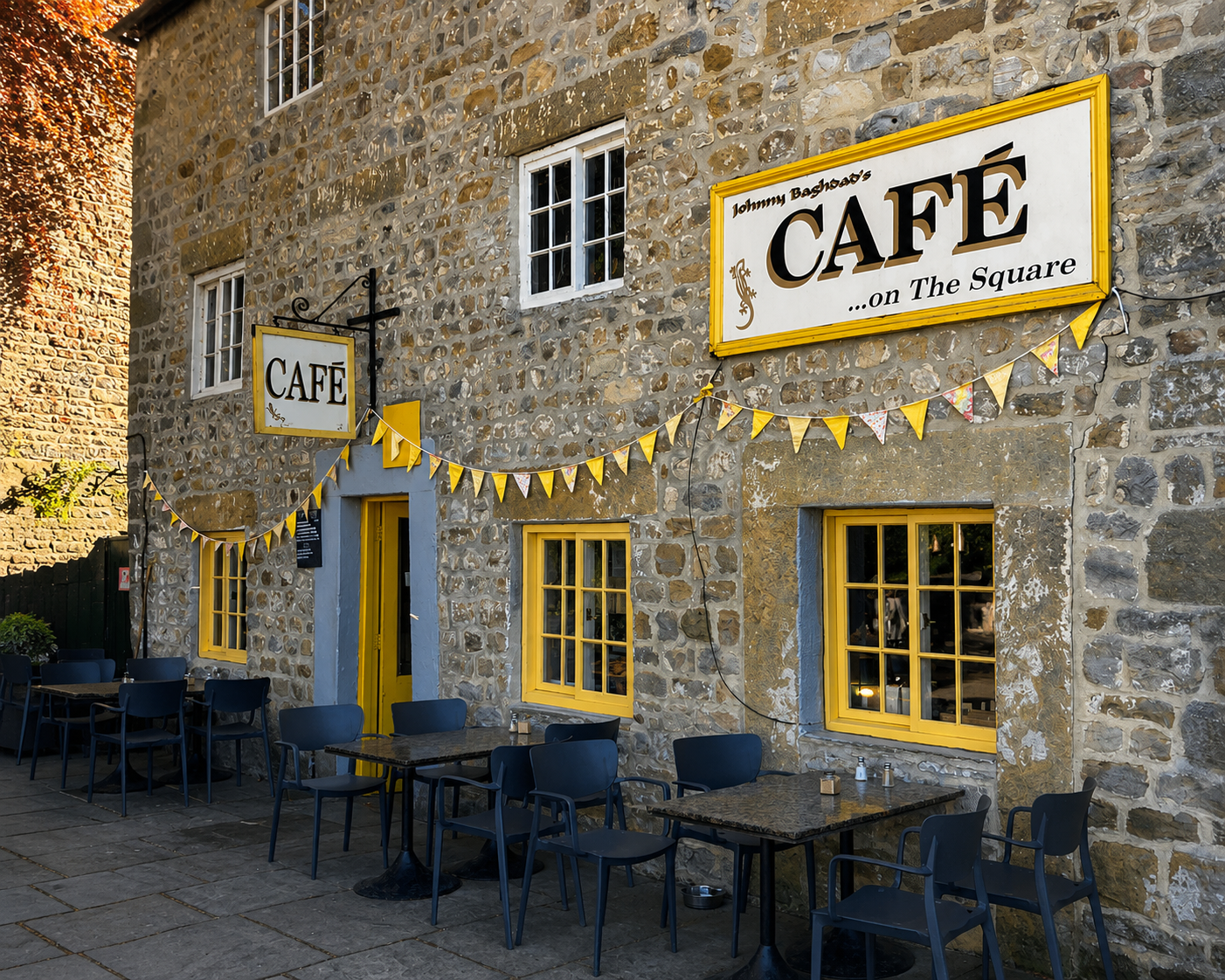 Exterior of a stone building with yellow-framed windows and doors, and signs reading 'Johnny Bashoot's Café on the Square'. Outdoor seating with black chairs and tables is arranged in front, decorated with yellow bunting.