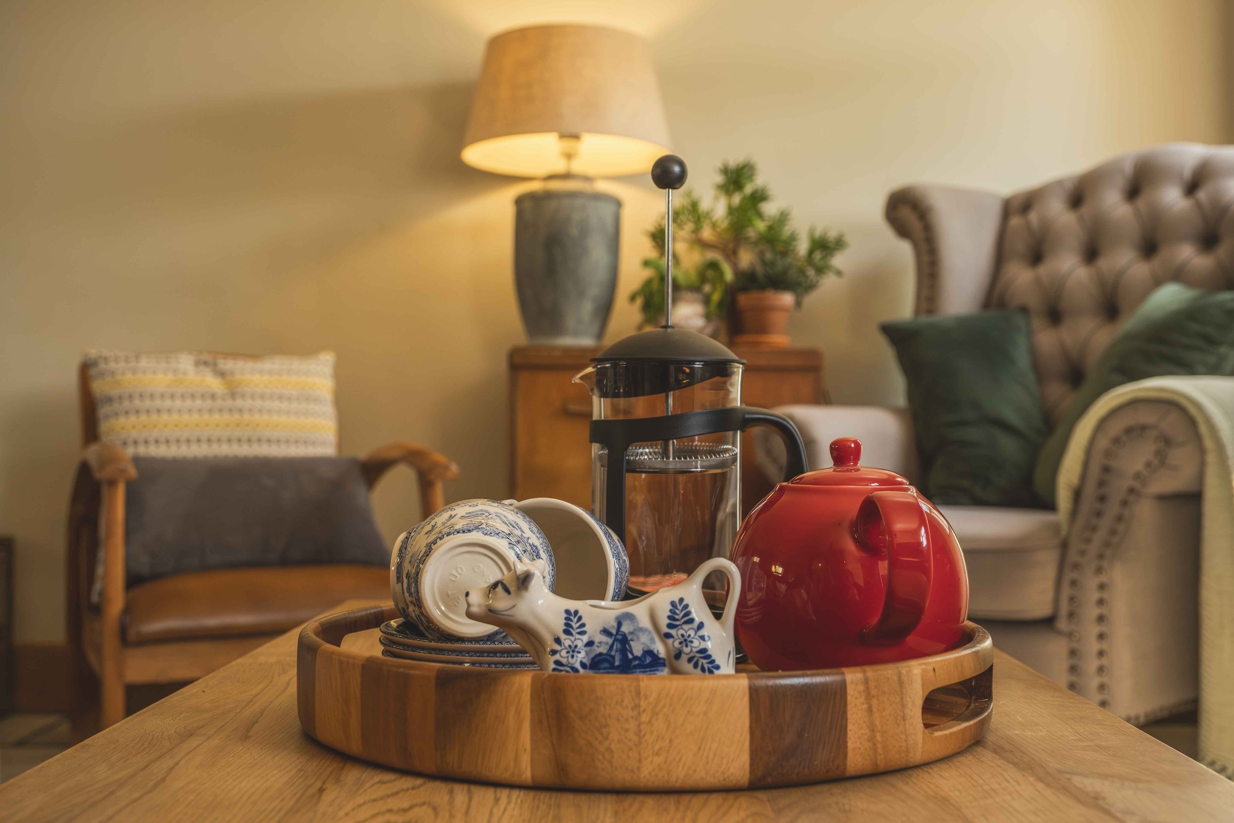 Close-up of a round wooden tray on a table with various teapots, cups, and mugs, with a living room in the background including a beige tufted armchair, a lamp, and a potted plant.