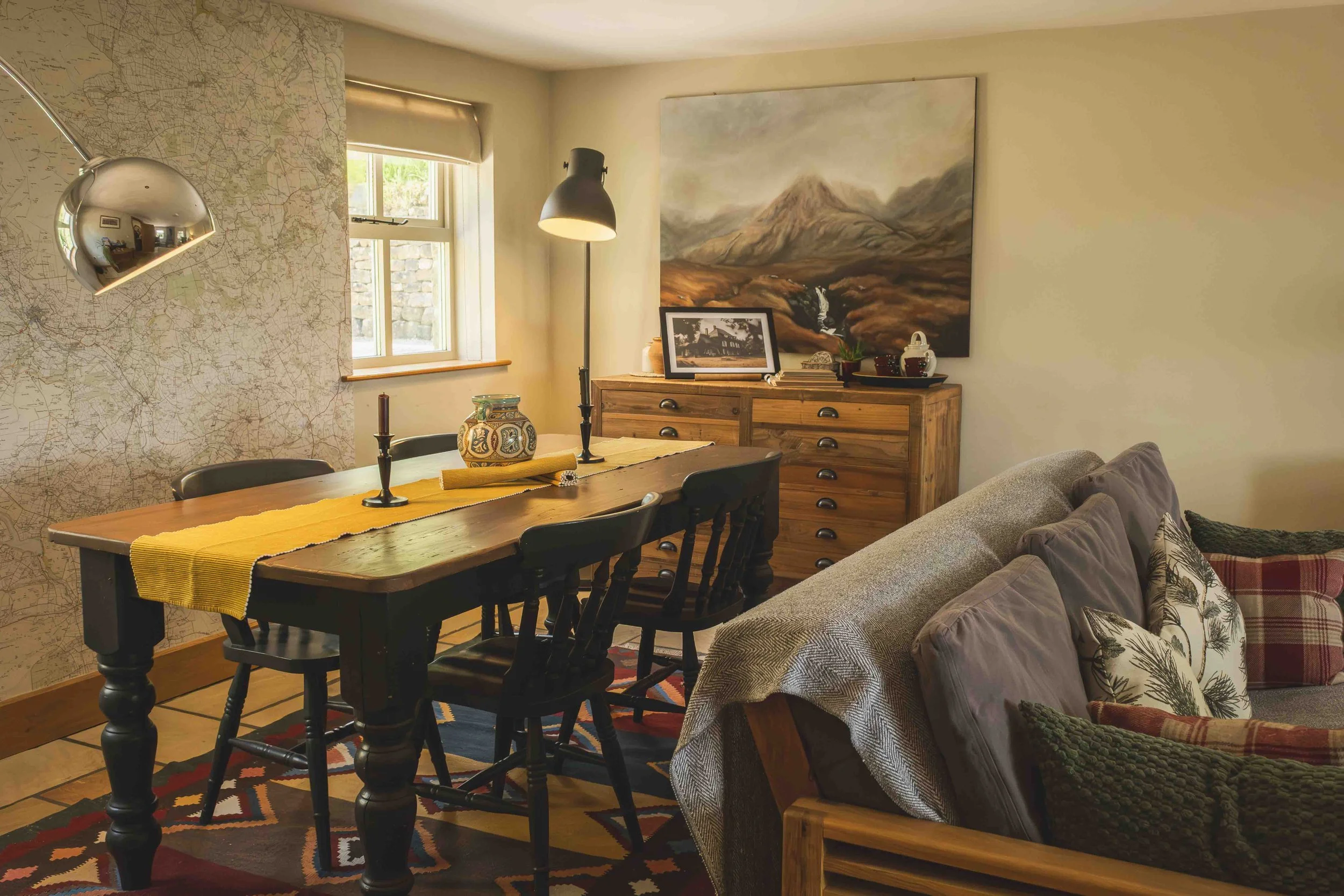 A cozy dining area with a wooden table covered with a yellow table runner, surrounded by black chairs, and lit by a black floor lamp. To the right, there is a beige couch with decorative pillows, and a large landscape painting of mountain scenery on the wall. The wall on the left has a large map or print, and a chrome wall lamp. A window with a beige shade is also visible.