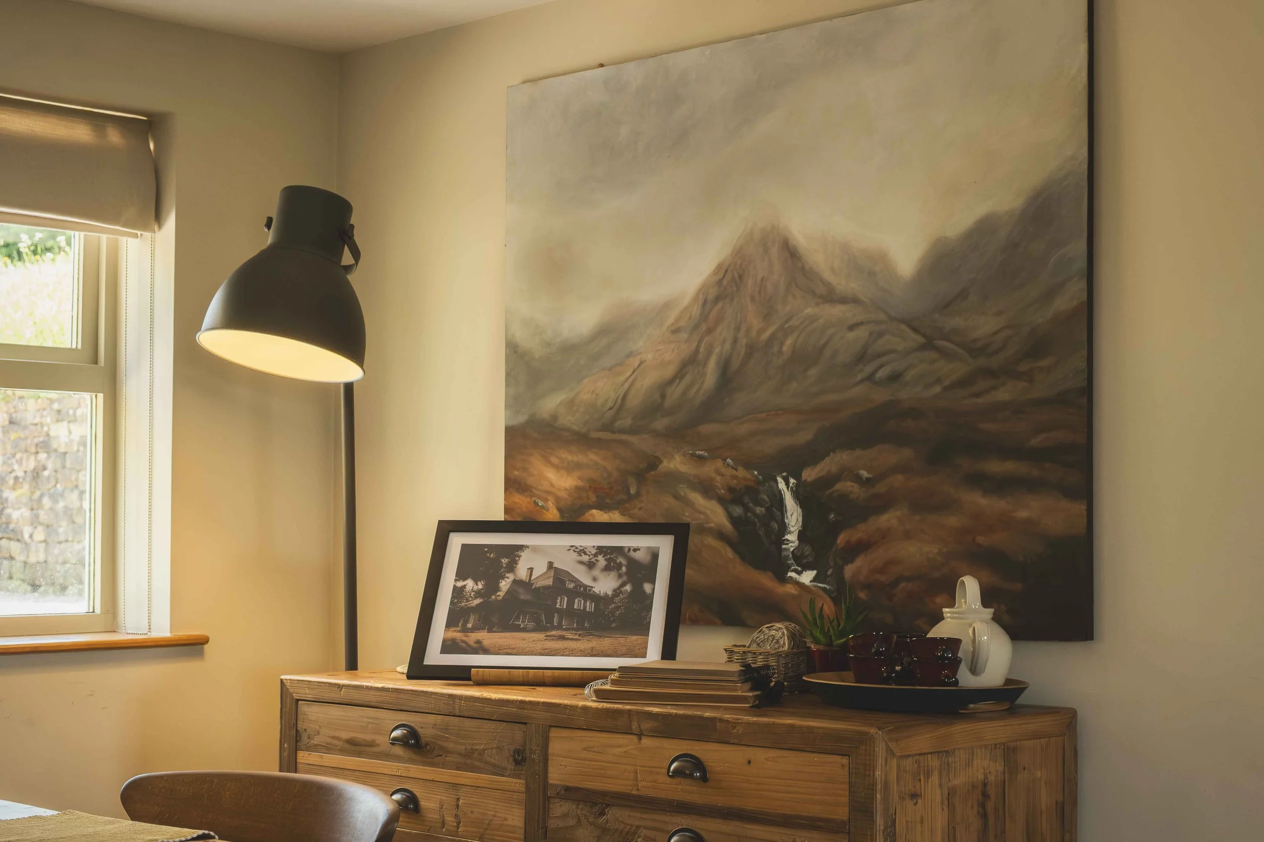 A corner of a room with a window, a wall-mounted black lamp, and a wooden sideboard with framed pictures, decorative vases, and plants. A large landscape painting of mountains hangs on the wall.