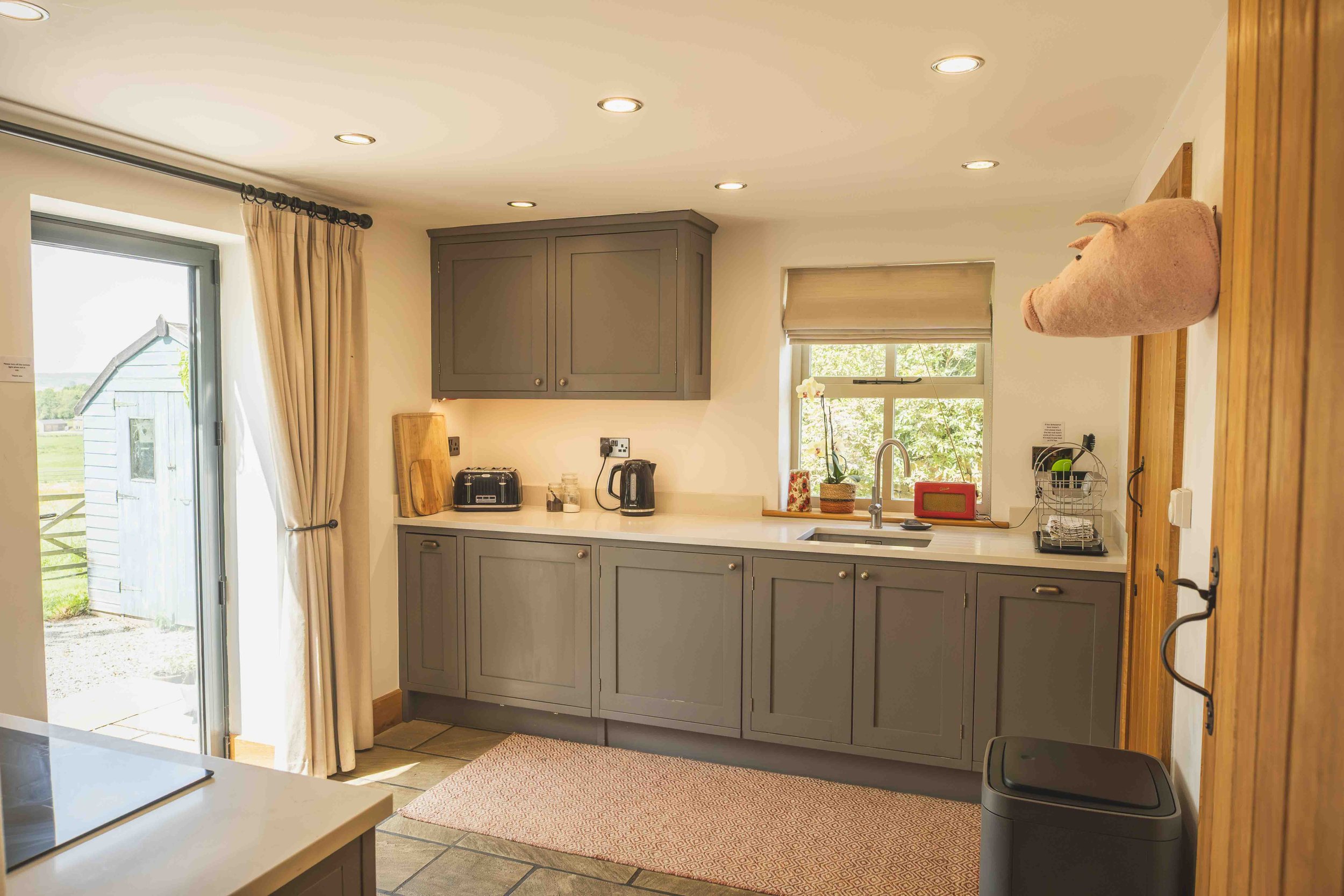 A kitchen with grey cabinets, a white countertop, and two windows. There is a toaster, kettle, and a cutting board on the counter. A pink plush pig-shaped pillow is mounted on a wooden door frame. The kitchen has recessed lighting and a small pink rug on the tiled floor.
