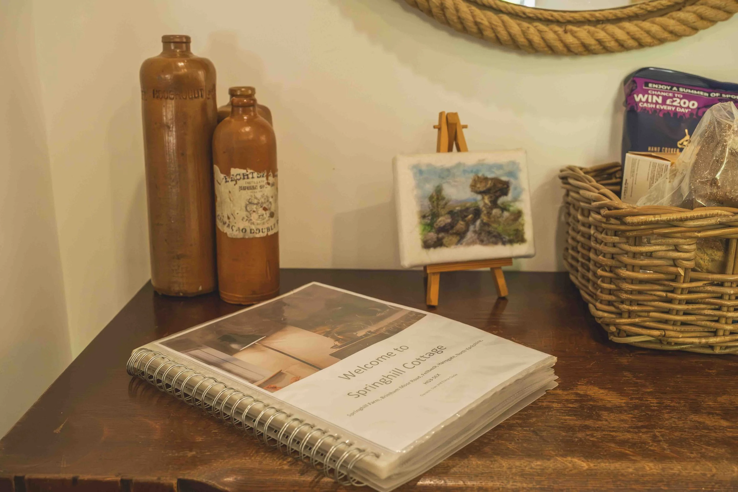 A desk with brown bottles, a small framed landscape painting, a spiral notebook with a pamphlet on top labeled "Welcome to Springhill Cottage," and a wicker basket, with a rope wall hanging above.