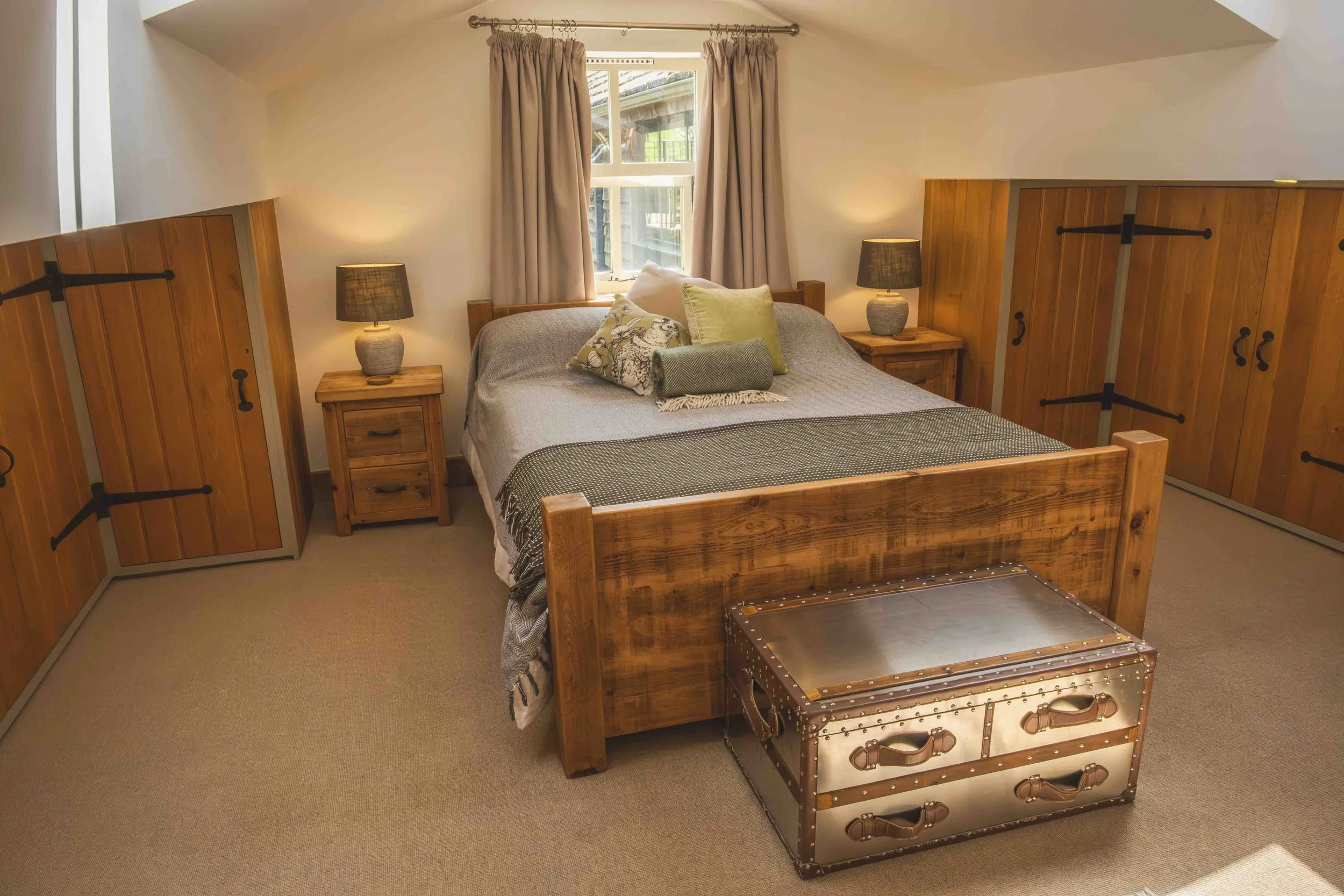 A cozy bedroom with a wooden bed frame, a gray bedspread, and multiple pillows. There are two nightstands with matching lamps and a window with beige curtains. A metallic trunk with leather handles is at the foot of the bed.