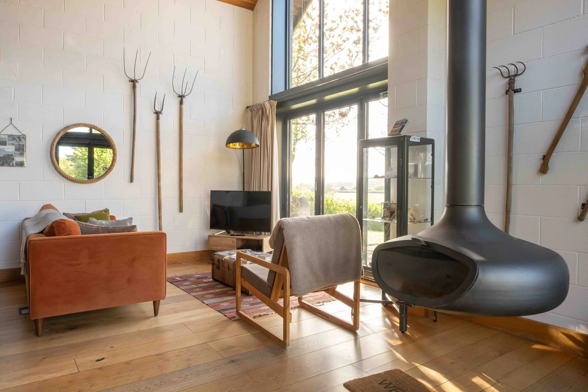Cozy living room with large window, orange armchair, gray chair, wooden floor, black wood stove, firewood box, and rustic gardening tools hanging on white brick wall.