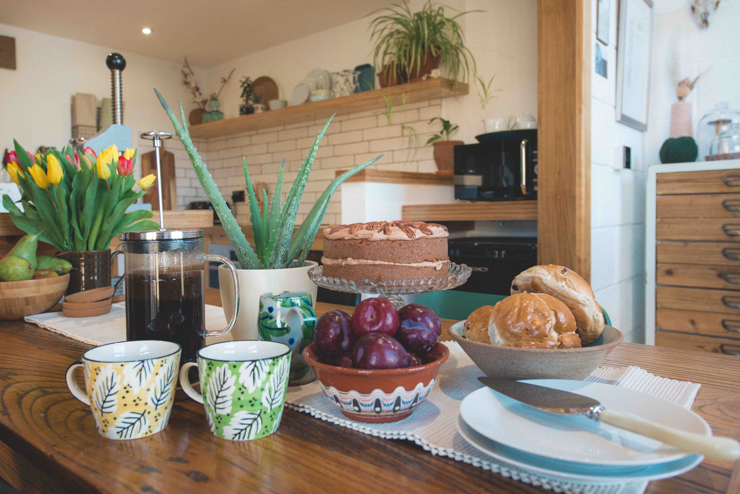 A cozy dining area with a wooden table set with colorful patterned mugs, a bowl of cherries, pastries, coffee, and a plant centerpiece. In the background, a kitchen with wooden shelves, potted plants, and a brick wall contributes to a warm, inviting atmosphere.