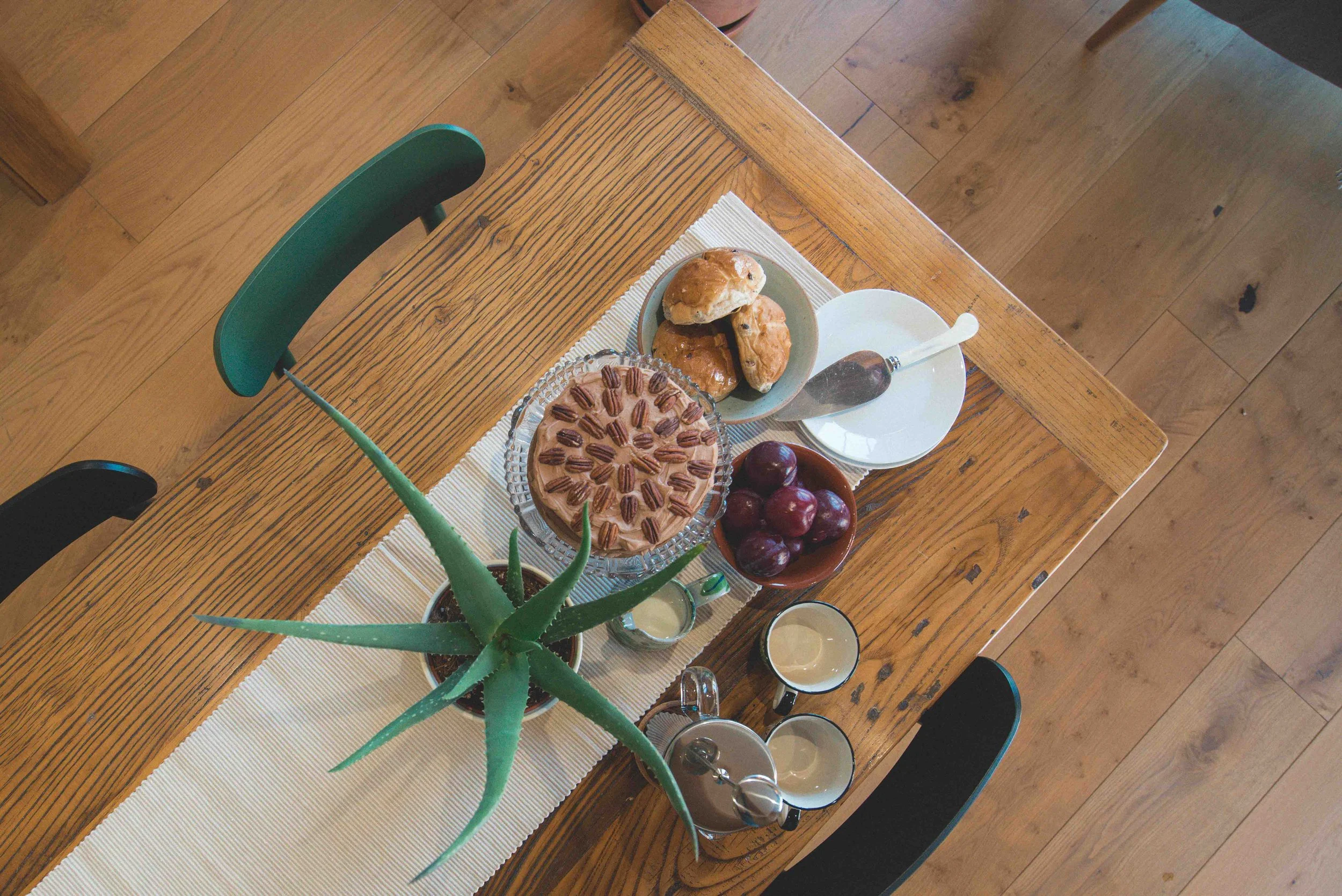 Top-down view of a wooden dining table with a beige table runner, decorated with a potted aloe vera plant, a pink pie with pecans, a bowl of cherries, a plate of croissants, and cups of coffee.