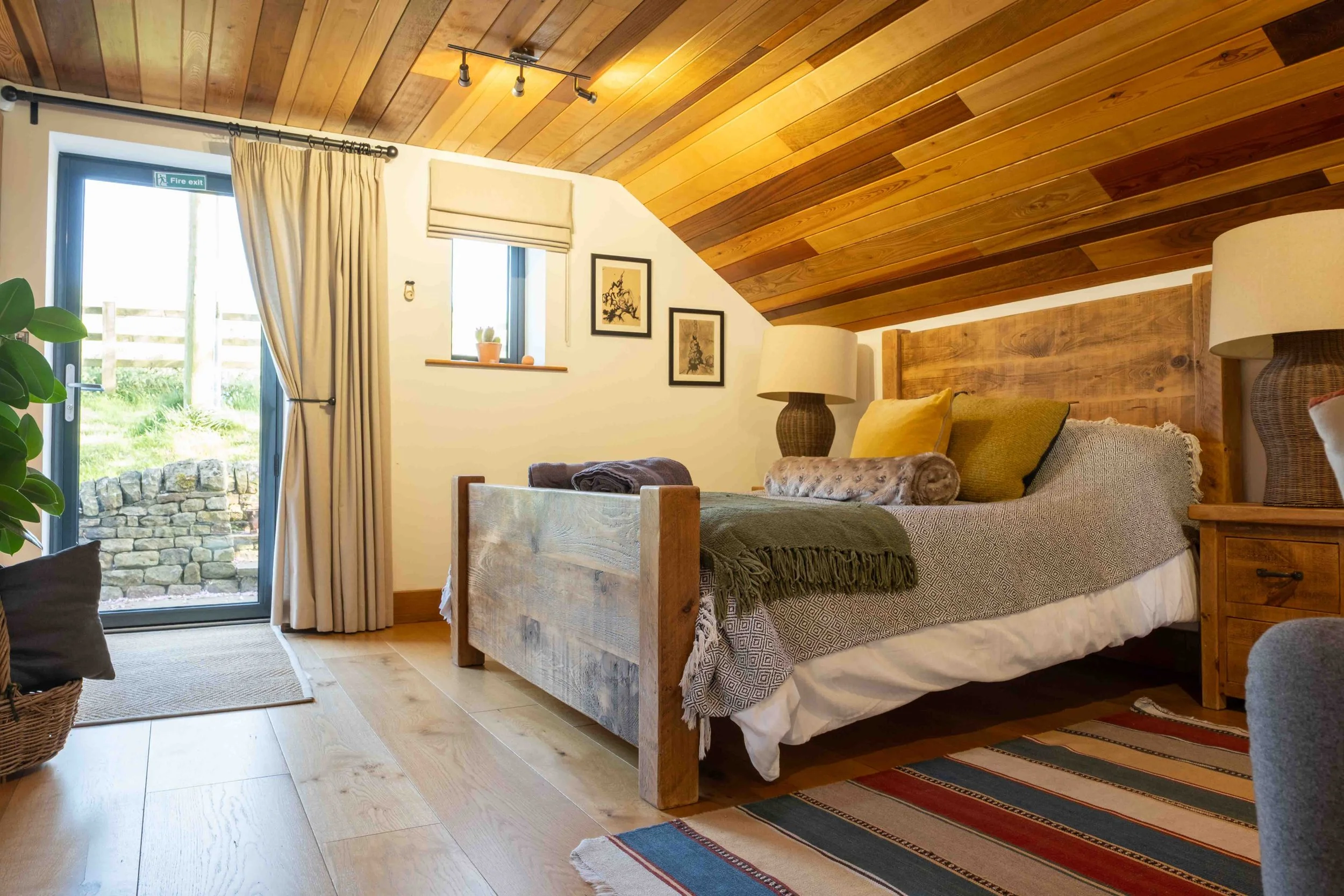 A cozy bedroom with a wooden sloped ceiling, beige curtains, a window with a plant and decorative items, paintings on the wall, a bed with yellow and beige pillows, a rustic bedside table with a lamp, a woven wicker chair, and a colorful striped rug on a wooden floor.