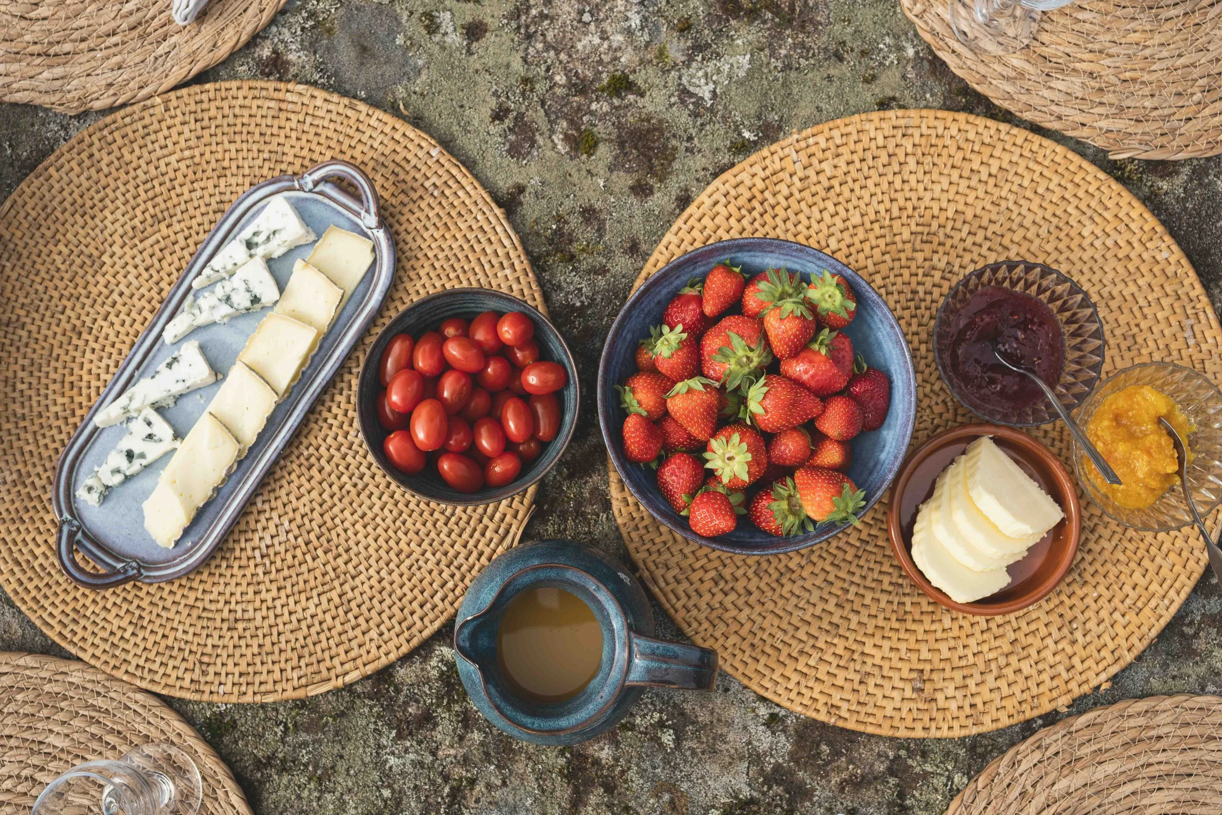 An outdoor breakfast spread with cheese, cherry tomatoes, strawberries, jam, butter, honey, and a cup of coffee on woven placemats.