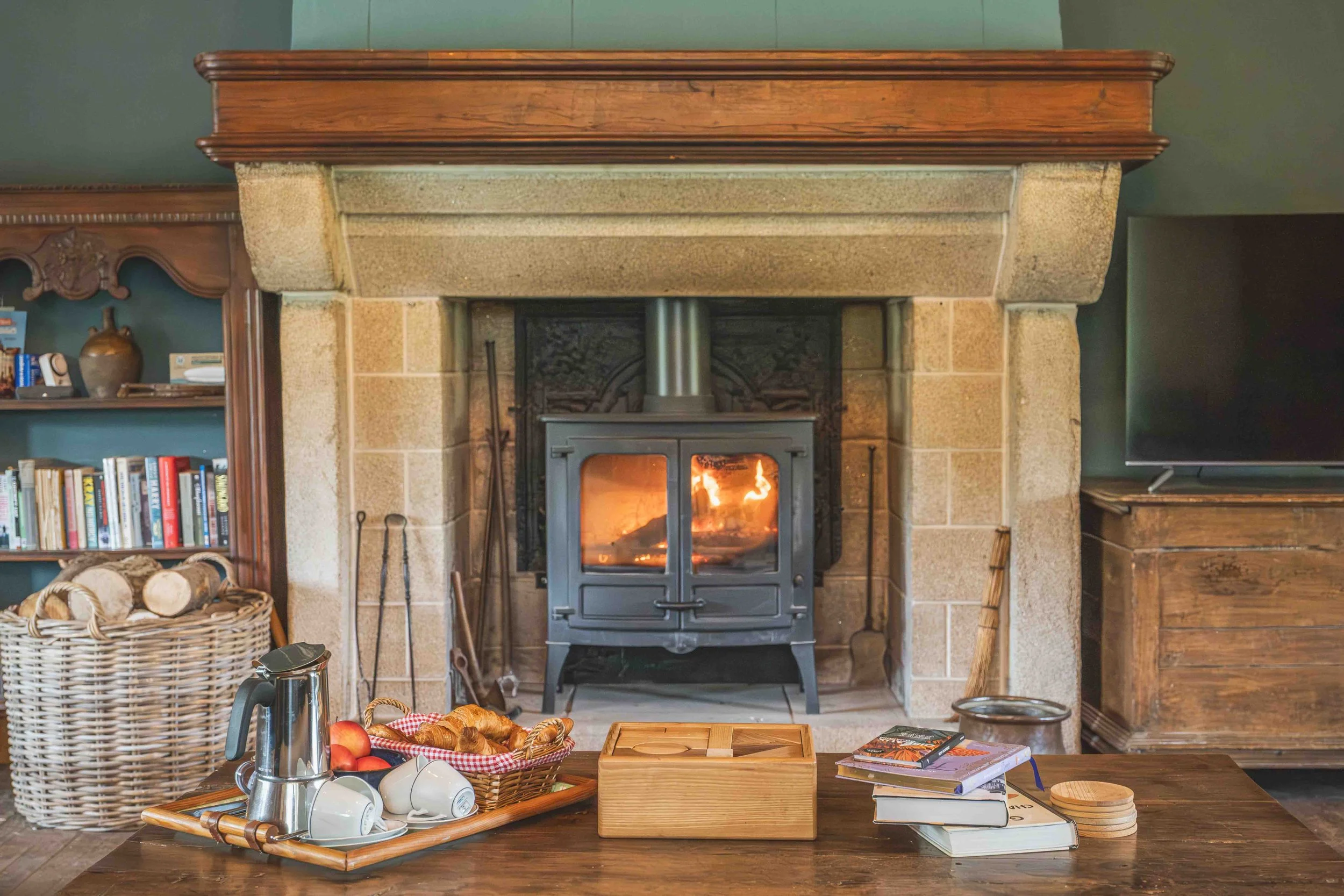 A cozy living room with a fireplace, bookshelves, and a table set with breakfast items including croissants, apples, mugs, and a kettle.