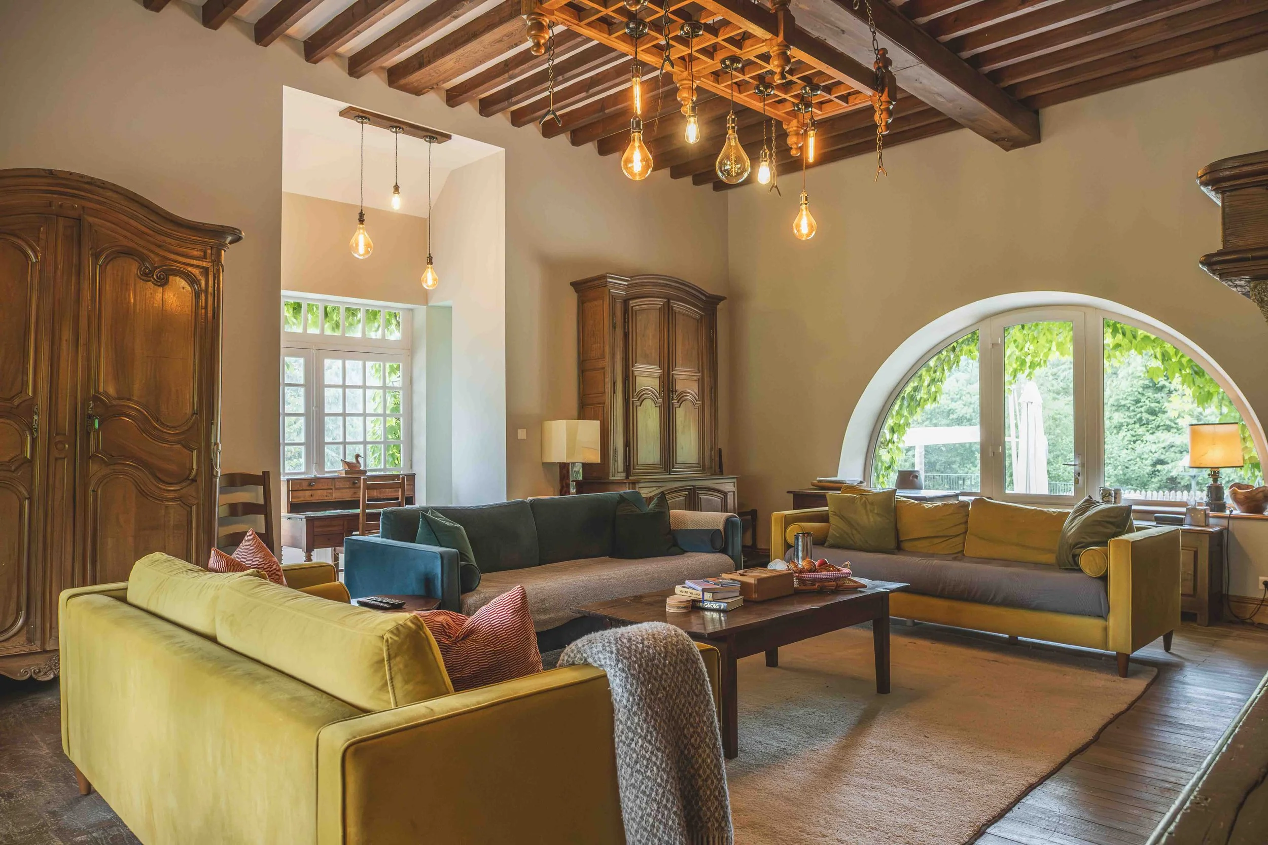 A spacious living room with a high wooden beam ceiling, a large window, and multiple pendant lights hanging from the ceiling. The room is furnished with a large yellow sofa, a green sofa, and a variety of other seating, with a wooden coffee table and various decorative items and books.