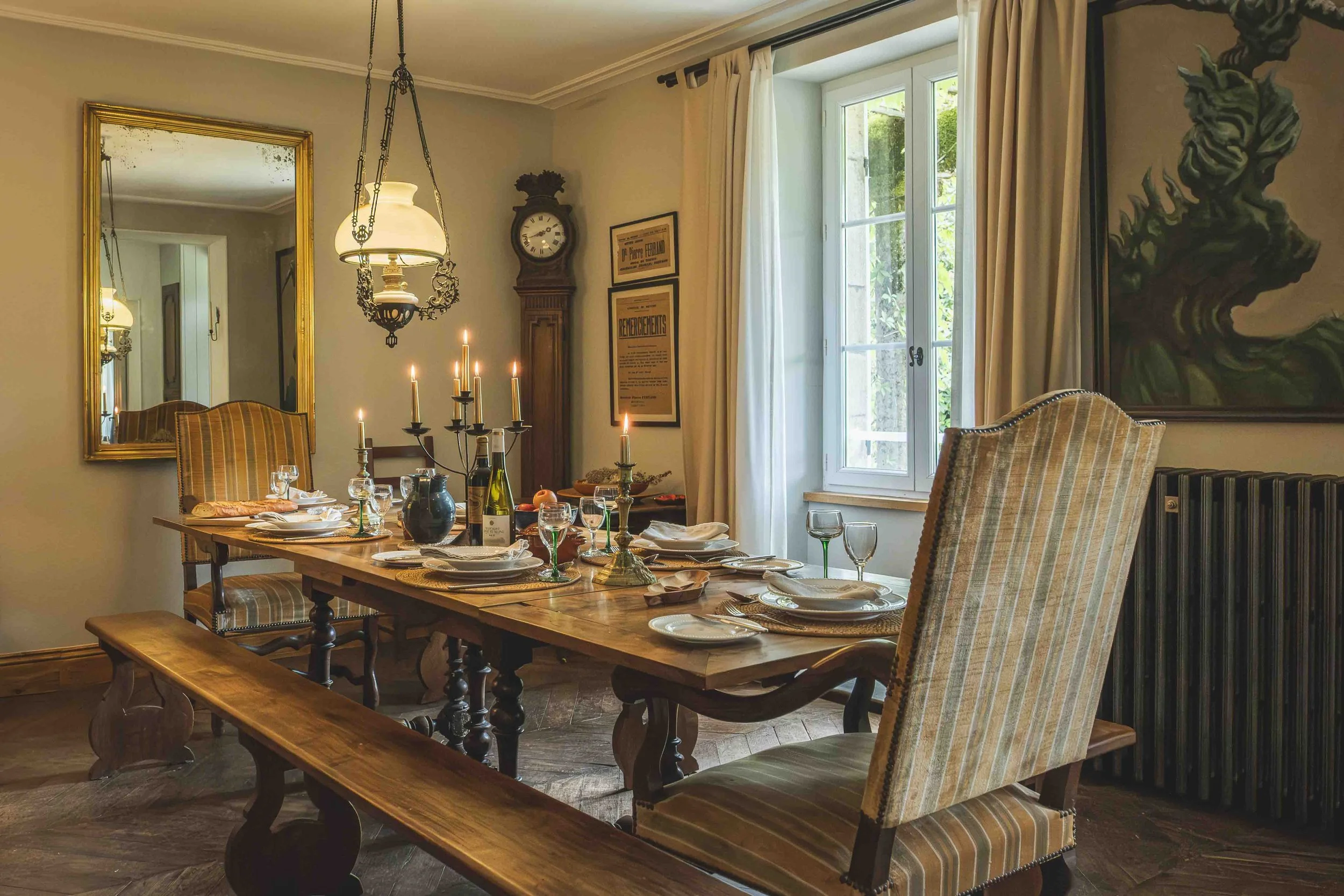 A dining room with a wooden table set for a meal, featuring wine bottles, glasses, plates, and candles. The room includes a large mirror, an antique clock, framed artwork, and a window with curtains, with sunlight coming in.