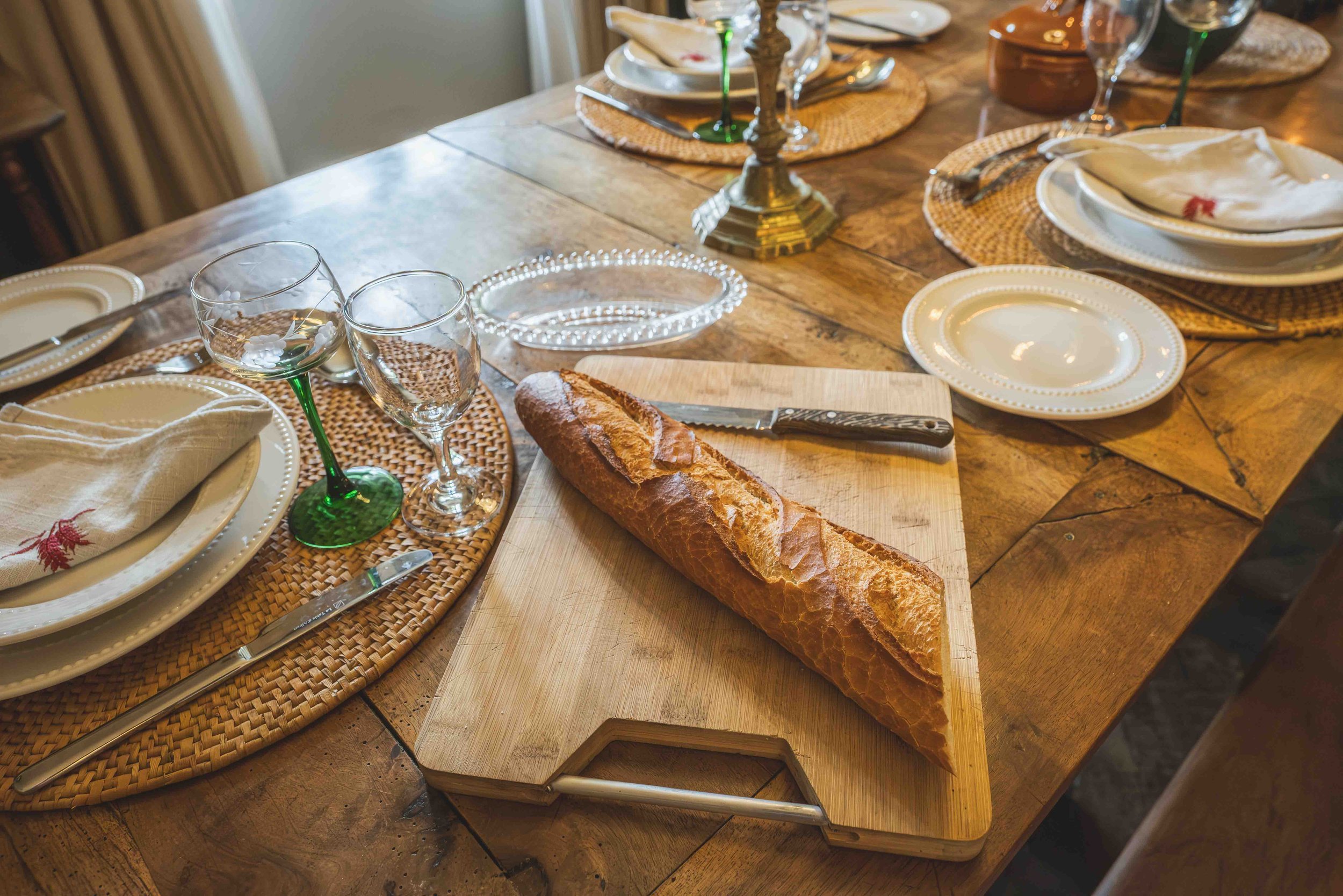 A rustic dining table set for a meal with plates, wine glasses, napkins, silverware, and a baguette on a cutting board.