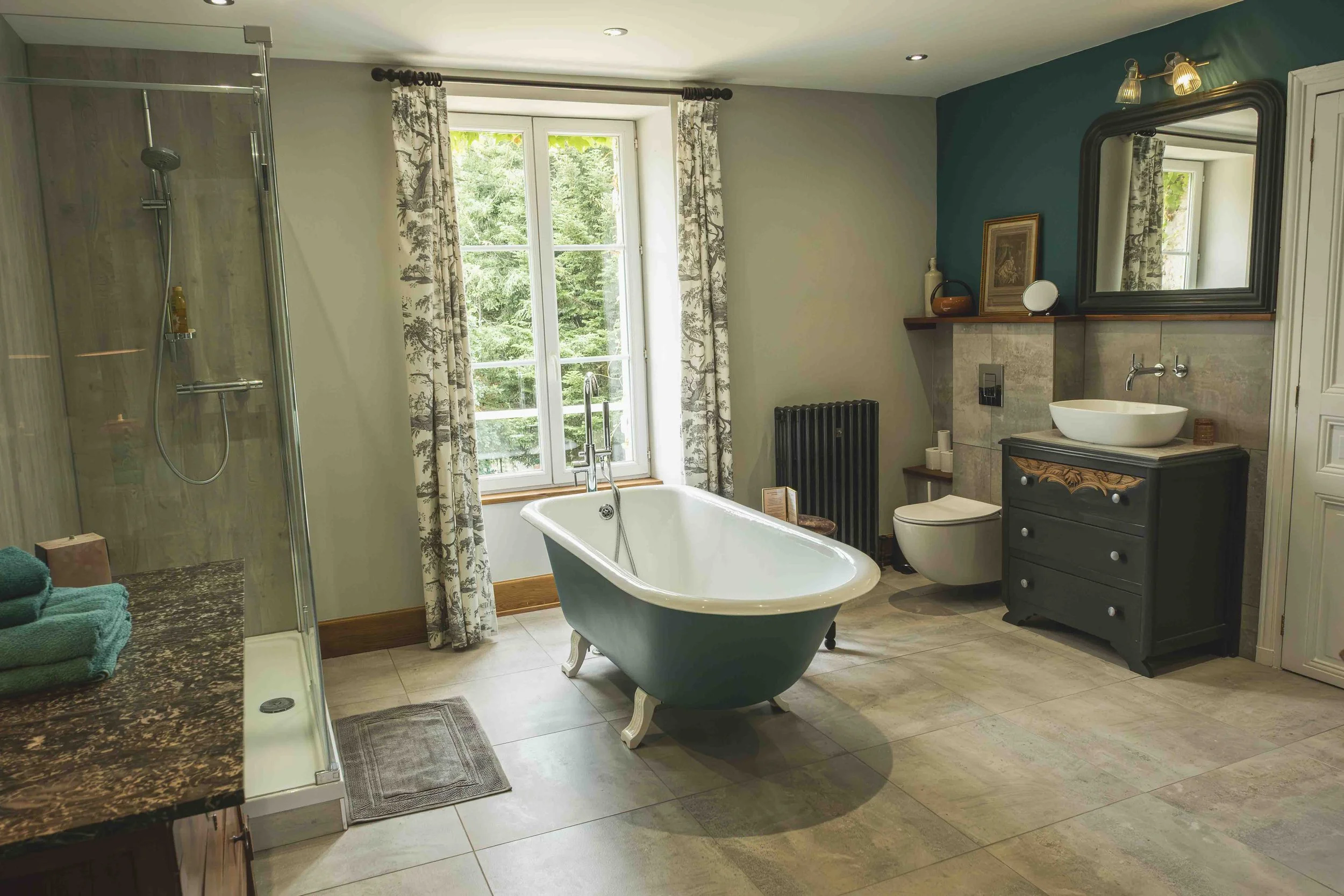 A spacious bathroom with a large window showing green trees outside, a vintage clawfoot bathtub in the center, a modern vanity with a round vessel sink on the right, a mirror above the sink, and a shower on the left with glass doors and a rainfall showerhead. The room features a mix of contemporary and classic decor, with beige and teal walls, and a radiator near the window.