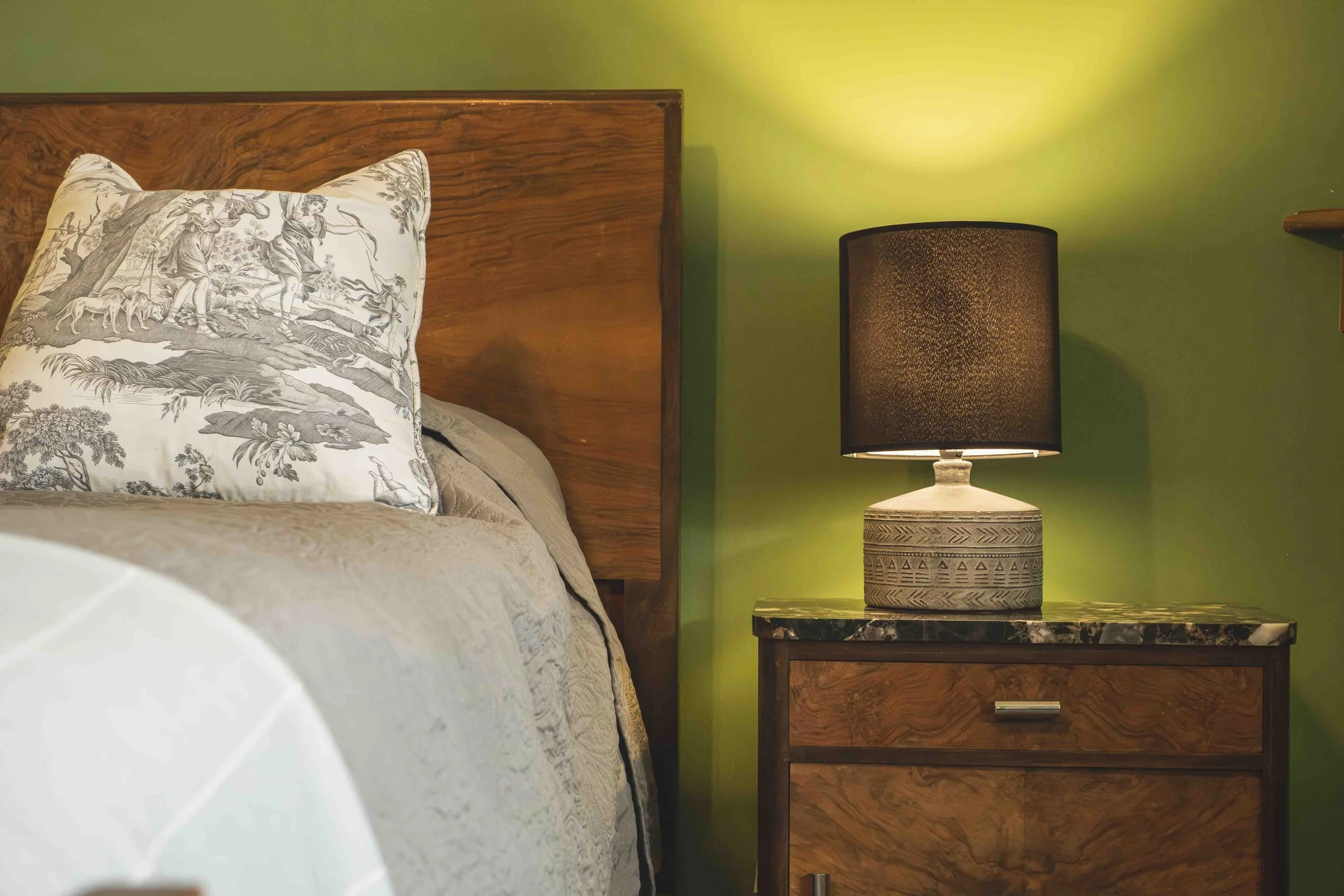 A bedroom with a wooden headboard, a patterned pillow, a bedside table, a black lampshade, and a green wall.