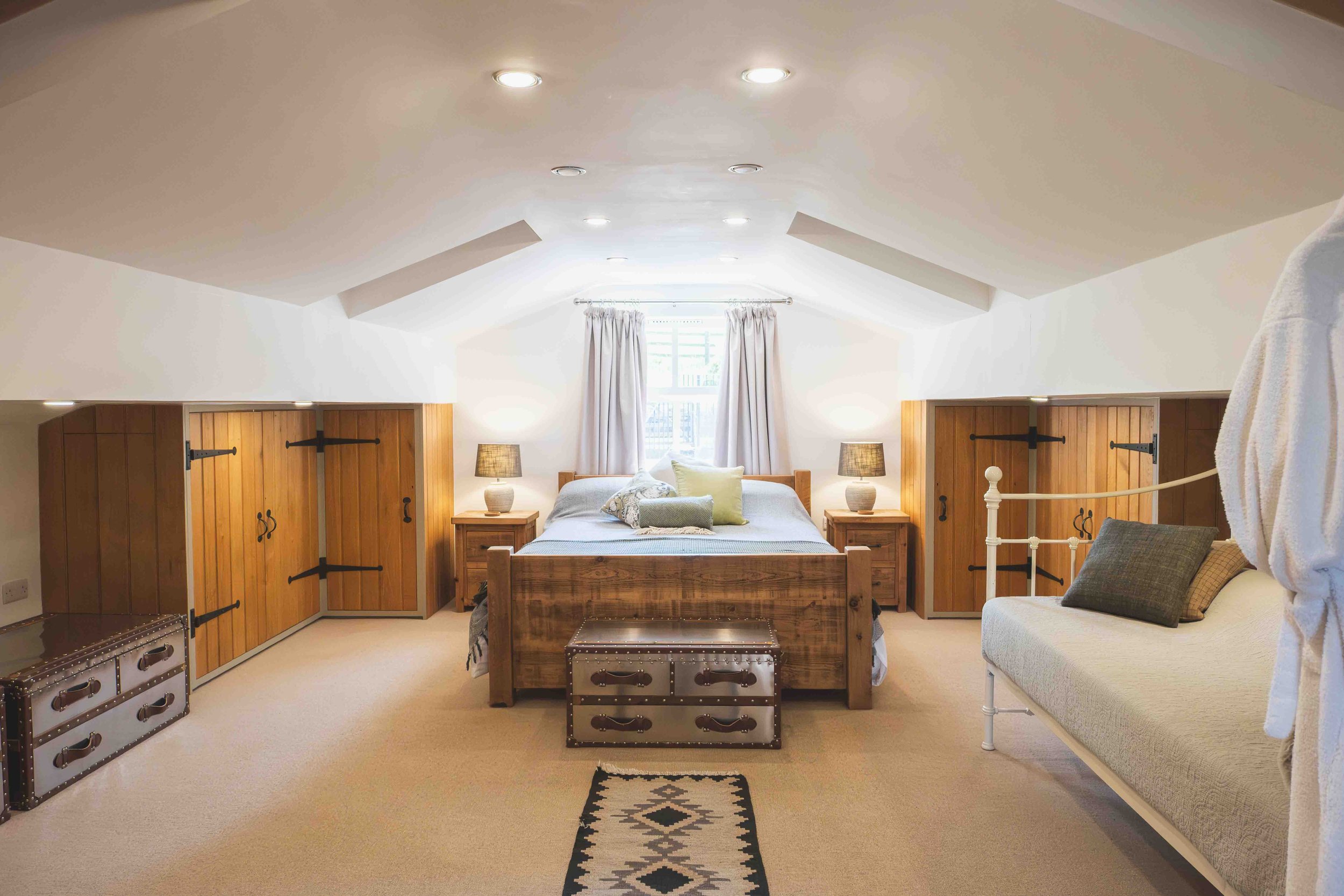 A cozy bedroom with a wooden bed frame, two matching nightstands with lamps, a window with curtains, and a sitting area with a sofa, pillows, and a blanket, illuminated by ceiling lights.