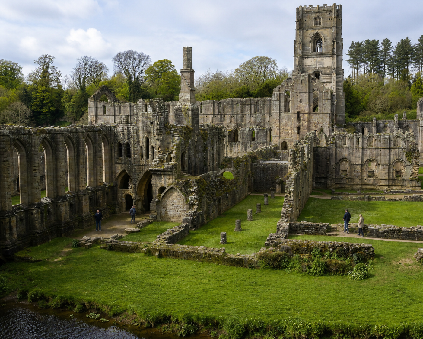 Ruins of an old castle with a large tower, surrounded by green trees and grass, with a small river in the foreground and a few visitors exploring the site.