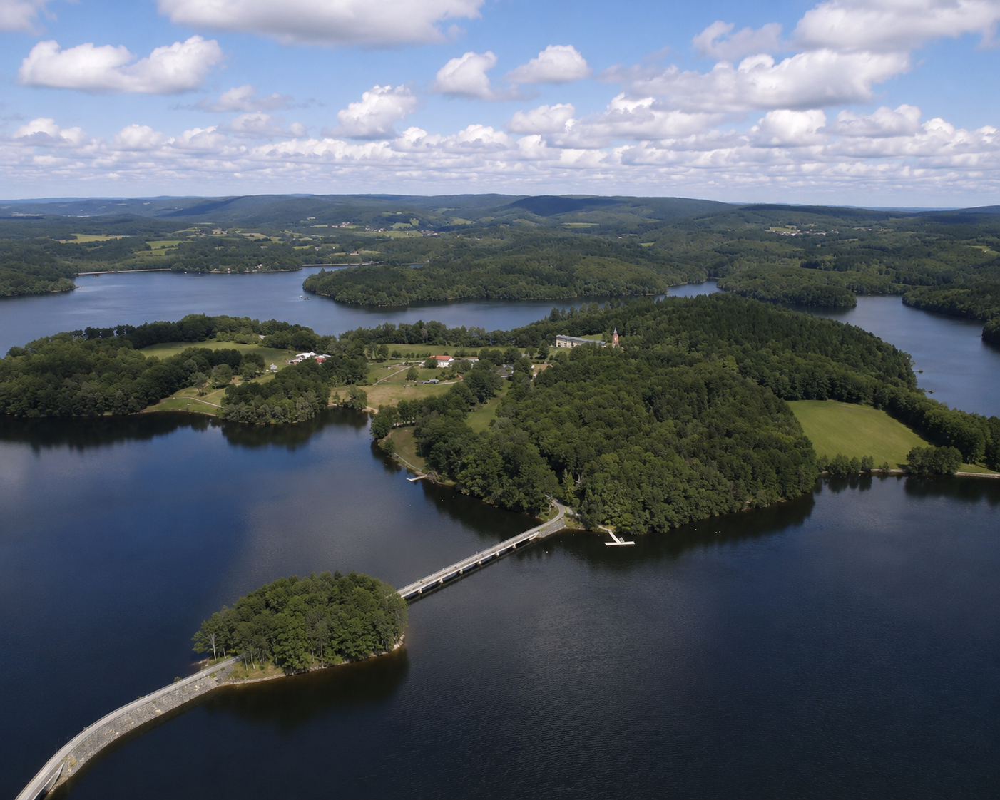 Aerial view of a scenic lake and surrounding green hills with a bridge connecting a small peninsula to the mainland, under a partly cloudy sky.