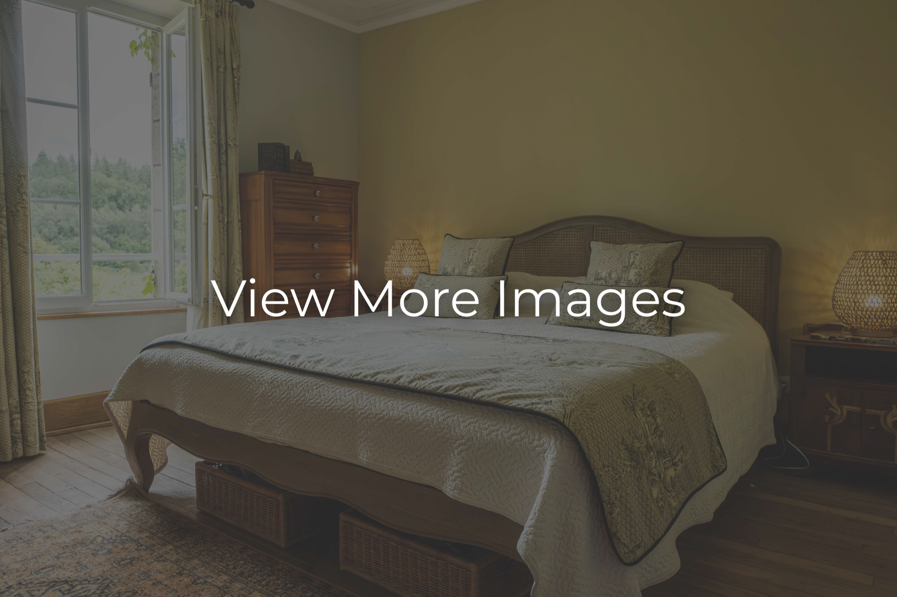 A cozy bedroom with a large bed, rattan headboard, beige bedding, decorative pillows, two bedside tables with lamps, a wooden dresser, and an open window with curtains revealing greenery outside.