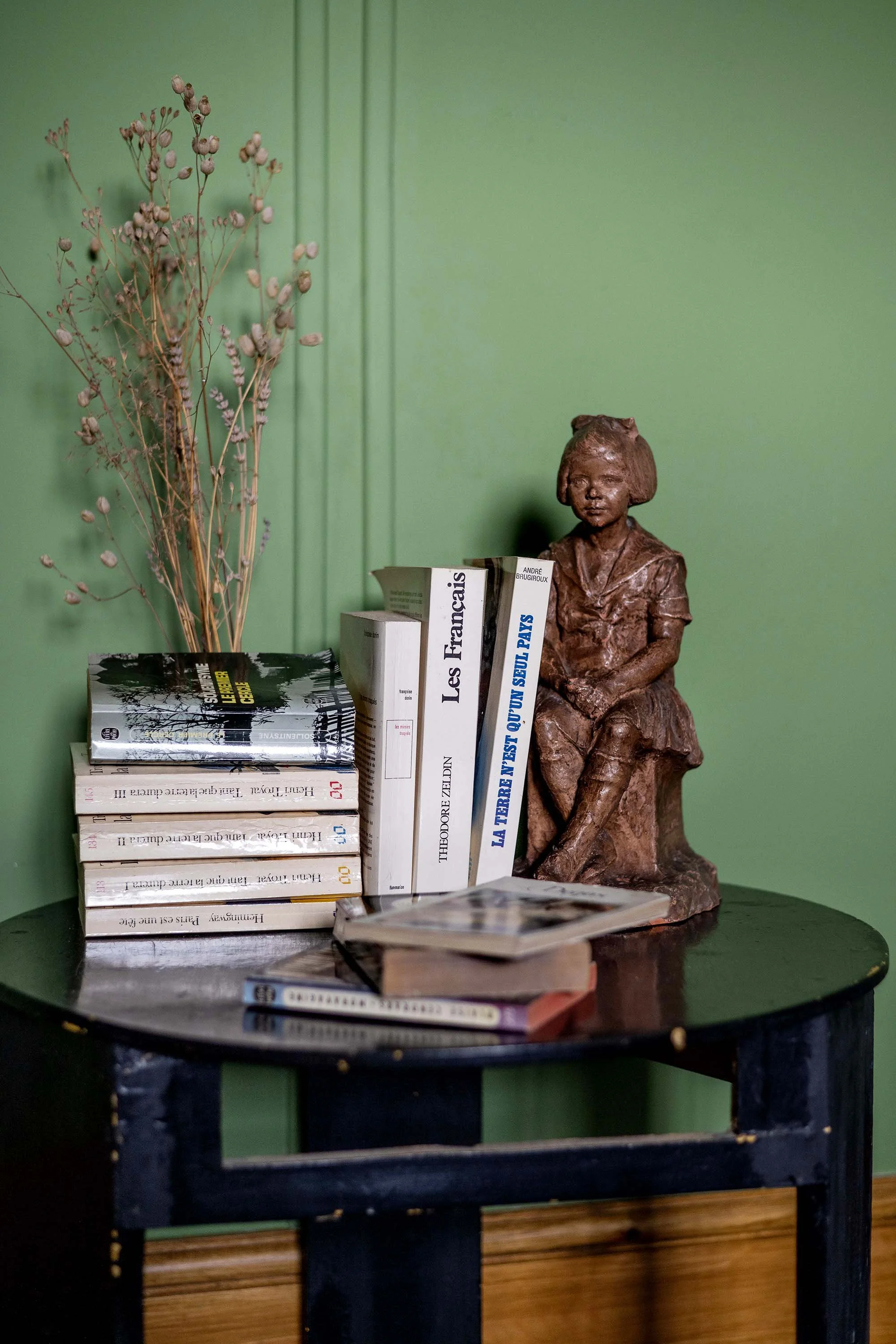 A small black table against a green wall. On the table is a sculpture of a woman sitting with crossed legs, made of bronze or similar material. To the left of the sculpture is a stack of books with mostly white covers, and one black-covered book on top. To the right of the sculpture are several books standing upright, with titles visible, including 'Les Francais' and others in French. In front of the books are two closed notebooks with a pen resting on them, and a string or cord around one of the notebooks.